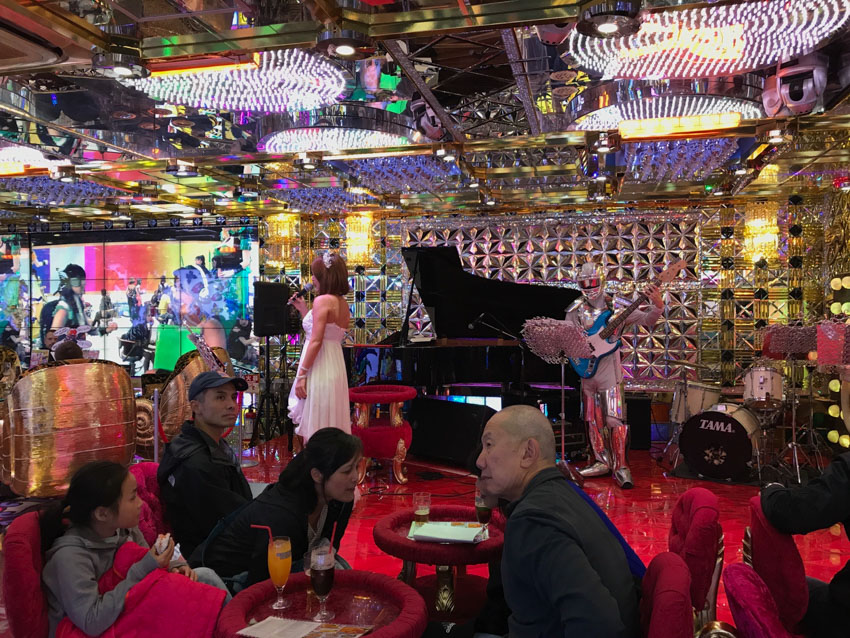 A woman in a white dress with a microphone on a stage, with her back slightly to the camera. A person dressed in a silver astronaut suit holds a blue electric guitar. There is a black grand piano in the background. Some people are sitting in front of the stage on pink cushion chairs. There is a reflective shiny wall behind the stage.