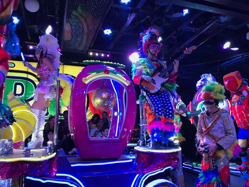 A float with glowing wires decorating it. There is a pink open cage on top of the float, in which a disco ball can be seen. Standing on the float and on either side of the pink cafe are a girl, not facing the camera, wearing a pink sequinned bikini top and bottoms, knee-high white boots and a white feather headpiece; and a man with pink glasses, blue pants patterned with white stars, a colourful top with colourful fluffy feathers, and is holding an electric guitar.