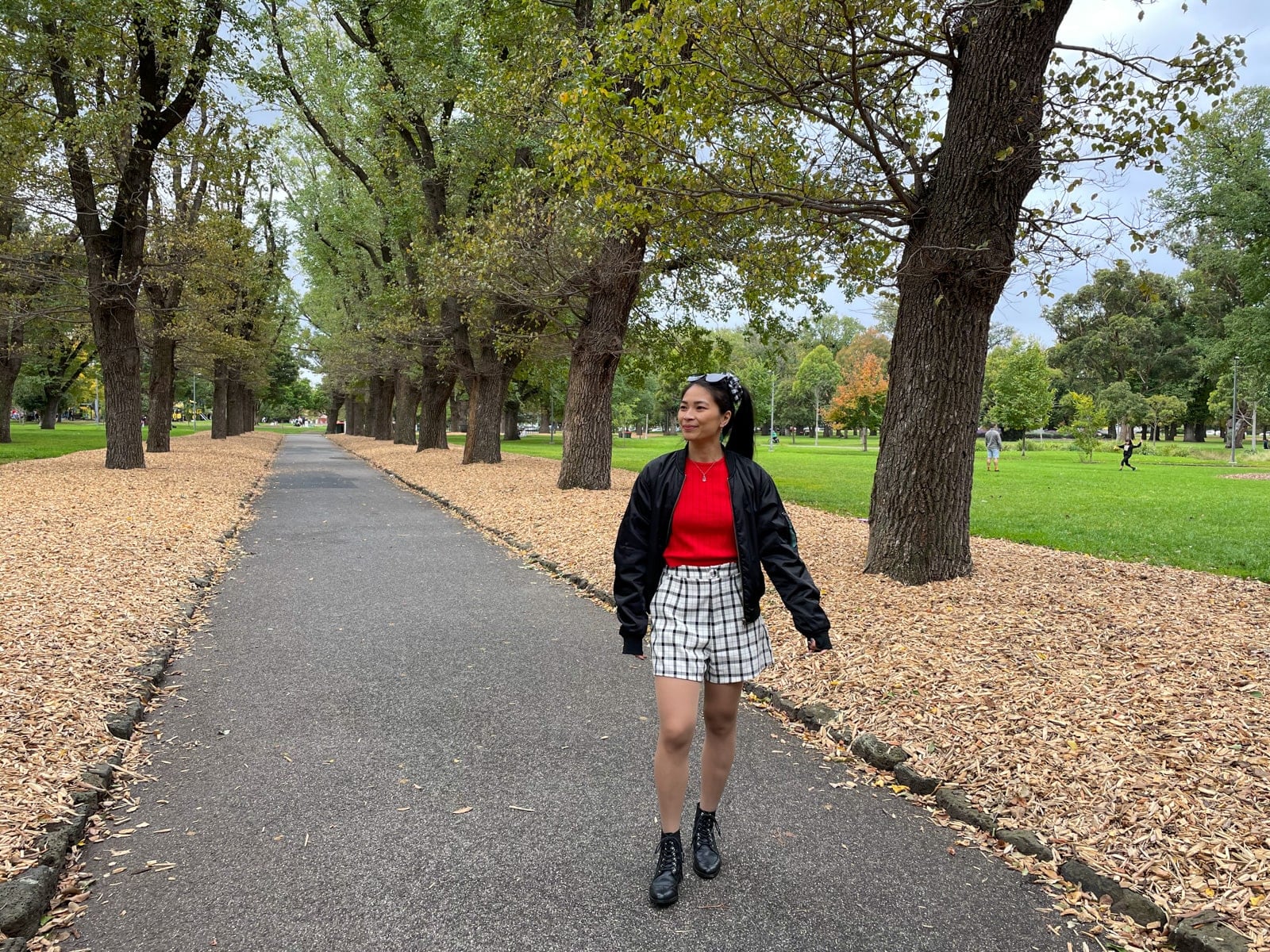 An Asian woman standing in a park with a concrete path going between planted trees. She has her long dark hair tied in a ponytail and a black-and-white gingham scrunchie. She is wearing a long sleeved red top with a black bomber jacket, black-and-white checkered shorts, and black lace-up boots.