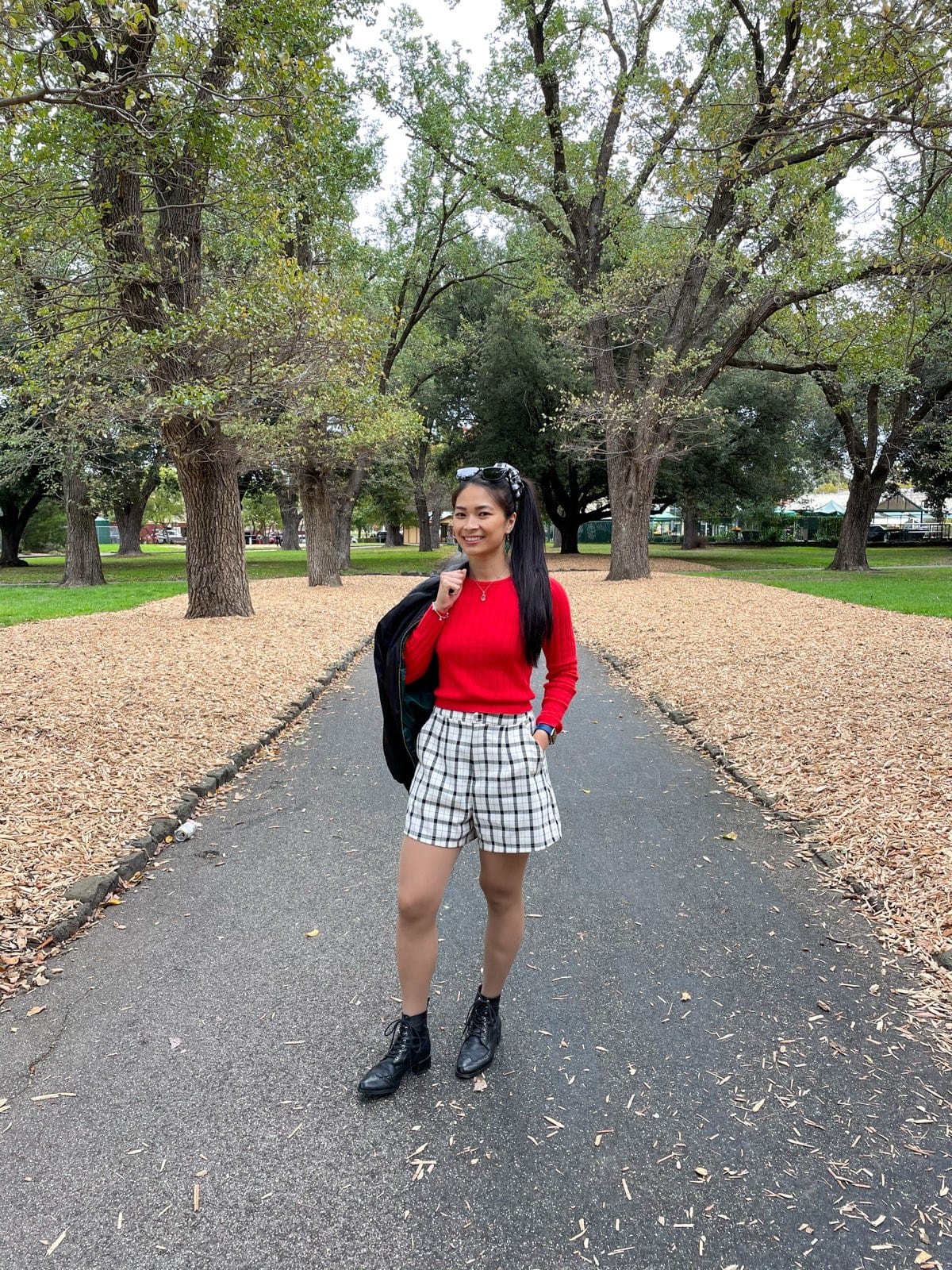 An Asian woman standing in a park with a concrete path going between planted trees. She has her long dark hair tied in a ponytail and a black-and-white gingham scrunchie. She is wearing a long sleeved red top, black-and-white checkered shorts, and black lace-up boots.