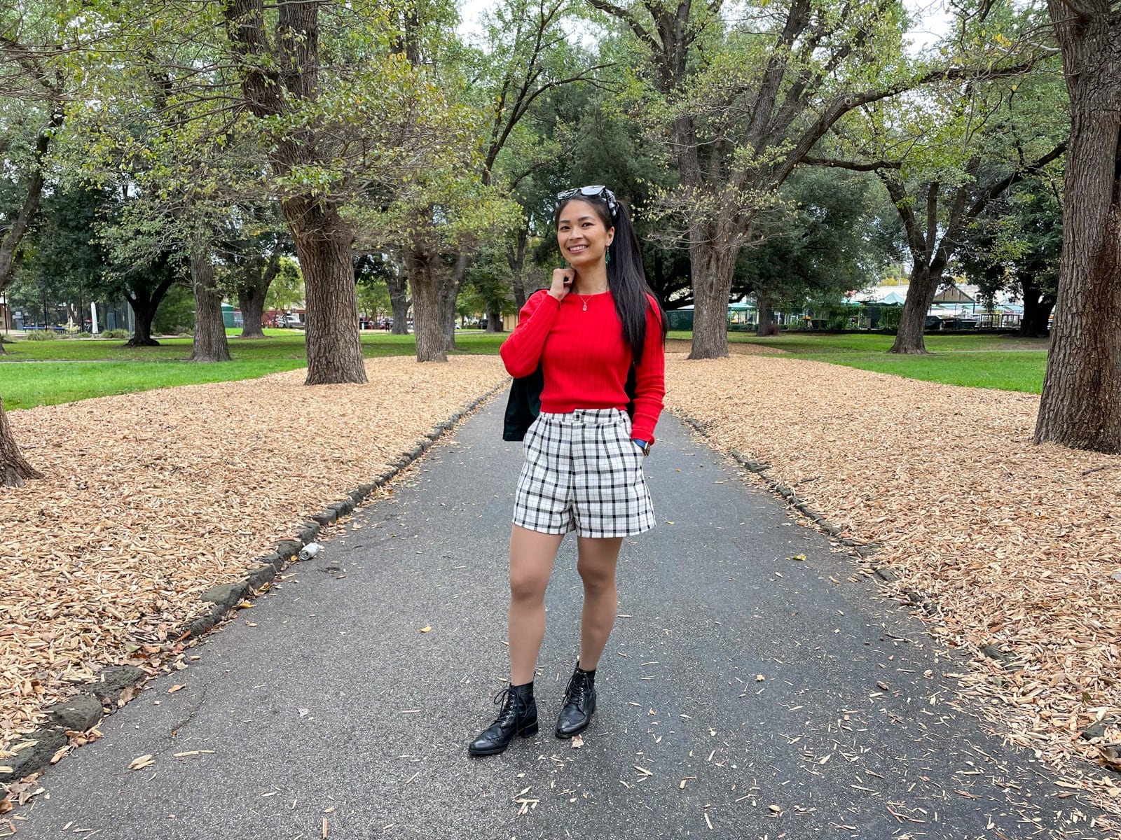 An Asian woman standing in a park with a concrete path going between planted trees. She has her long dark hair tied in a ponytail and a black-and-white gingham scrunchie. She is wearing a long sleeved red top with a black bomber jacket, black-and-white checkered shorts, and black lace-up boots.