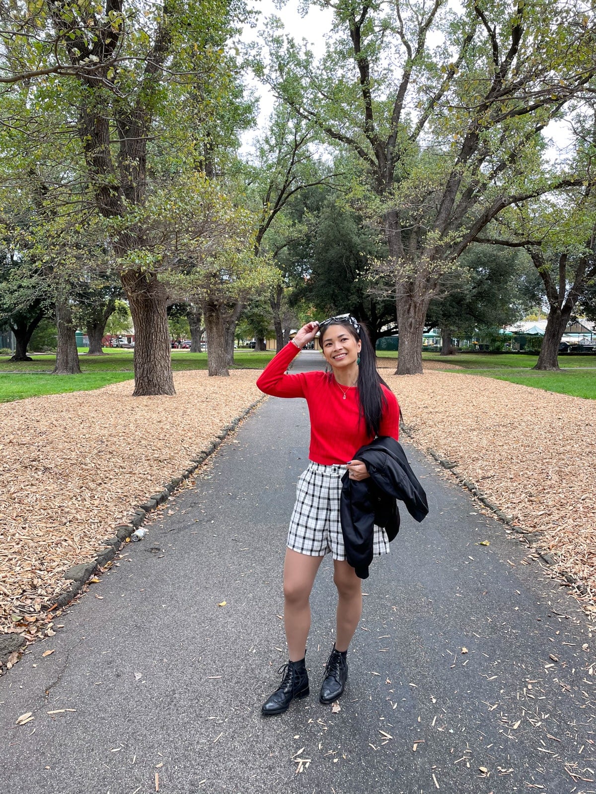 An Asian woman standing in a park with a concrete path going between planted trees. She has her long dark hair tied in a ponytail and a black-and-white gingham scrunchie. She is wearing a long sleeved red top with a black bomber jacket, black-and-white checkered shorts, and black lace-up boots. She is carrying a black jacket over one arm, and has one hand holding up the black sunglasses on top of her head.