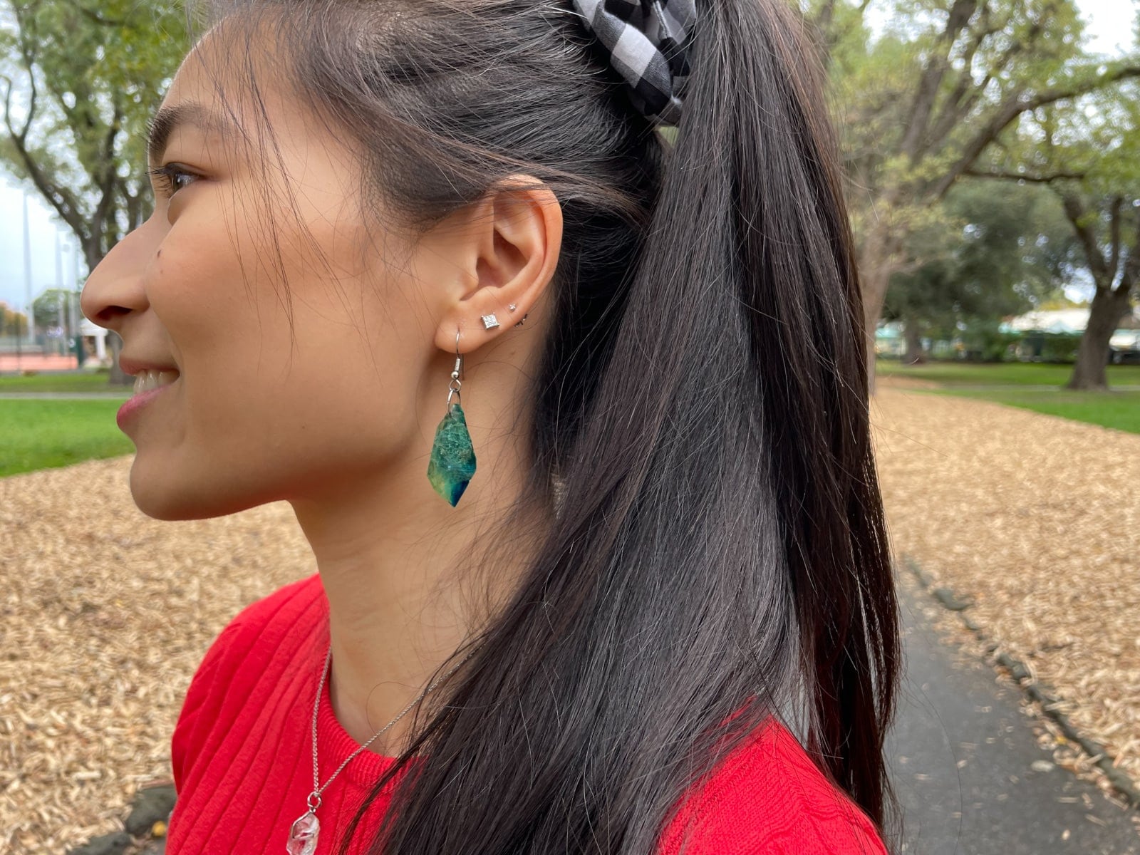 A close-up of the side of a woman’s face, showing her ear. She has a hook earring with a bright blue diamond-shaped resin drop in her lowest lobe piercing, and silver studs in other lobe piercings.