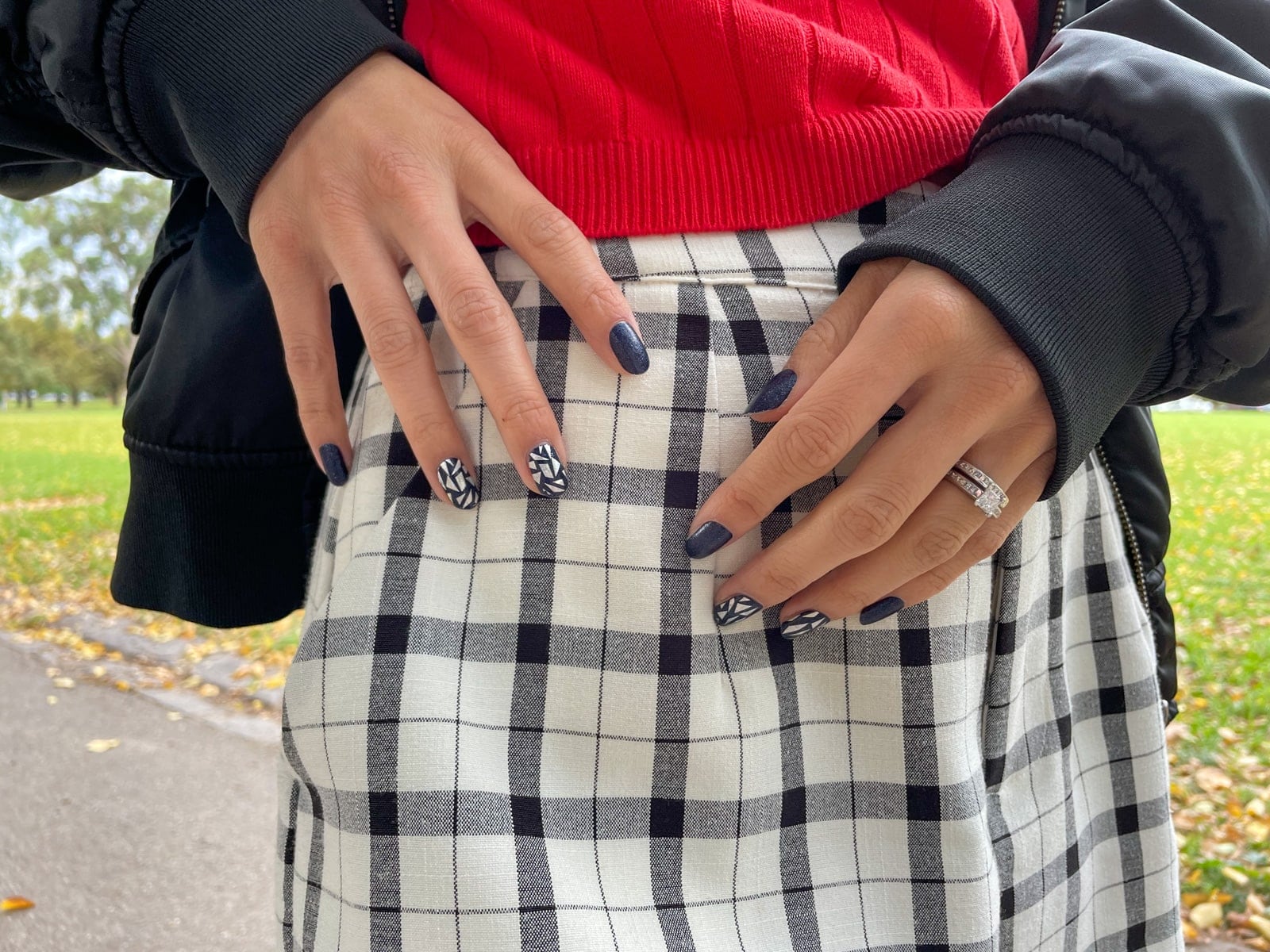 A close-up of a woman’s nail art, which is dark navy and slightly glittery, with some accent nails with silver geometric shapes. In the background is the woman’s black-and-white checkered shorts, and black lace-up boots.
