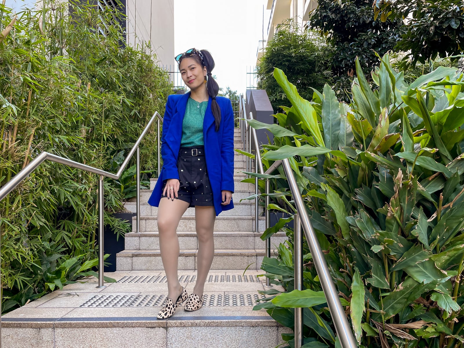 An Asian woman with dark hair tied in a ponytail with tied sections, standing on a set of concrete stairs. She is wearing a bright blue coat, a green top and black shorts, and giraffe print loafers. She has sunglasses on top of her head.