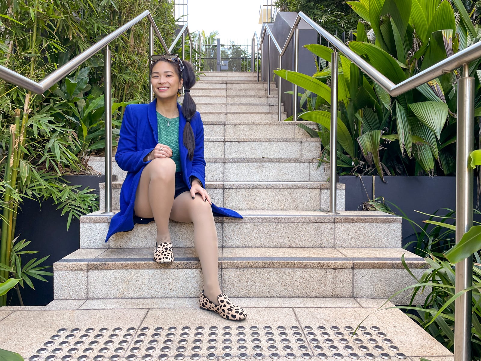 An Asian woman with dark hair tied in a ponytail with tied sections, sitting on a set of concrete stairs. Both her knees are bent with her hands relaxing on her knees. She is facing slightly towards the camera. She is wearing a bright blue coat, a green top and black shorts, and giraffe print loafers. She has sunglasses on top of her head.