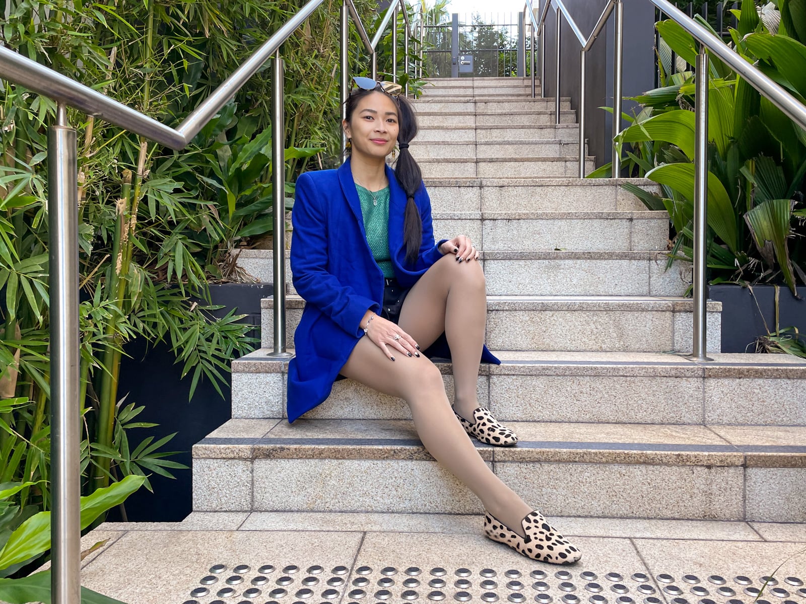 An Asian woman with dark hair tied in a ponytail with tied sections, sitting on a set of concrete stairs. One of her knees is bent and the other leg is outstretched with her hands relaxing on her knees. She is wearing a bright blue coat, a green top and black shorts, and giraffe print loafers. She has sunglasses on top of her head.