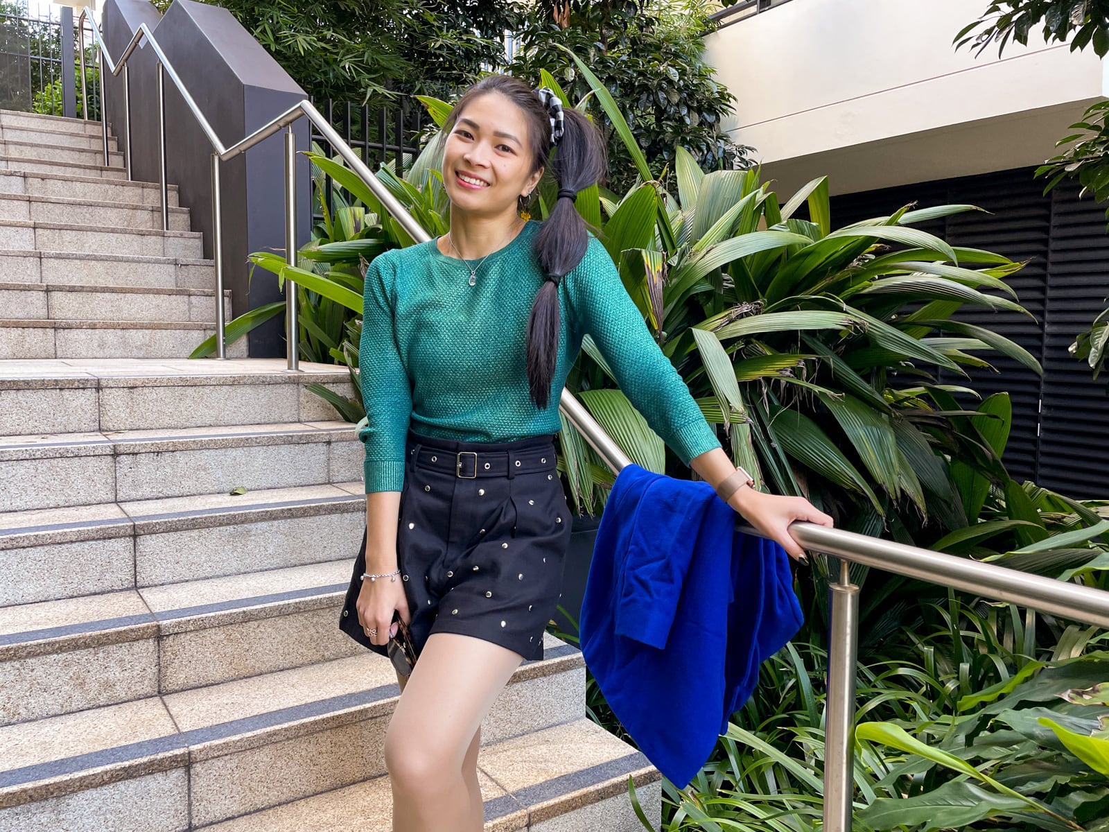 An Asian woman with dark hair tied in a ponytail with tied sections, standing on a set of concrete stairs. She is wearing a green top with three-quarter sleeves, and black shorts, and has her hand on a metal railing.