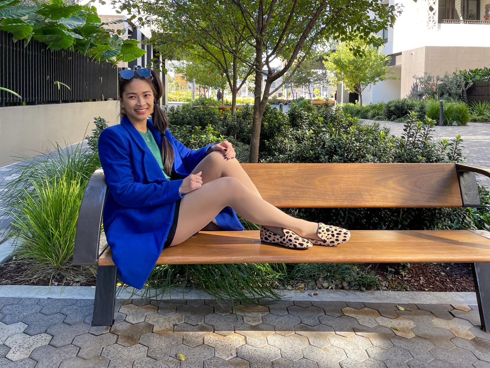 An Asian woman with dark hair tied in a ponytail with tied sections, sitting on a wooden bench sideways. She is wearing a bright blue coat, a green top and black shorts, and giraffe print loafers. She has sunglasses on top of her head.