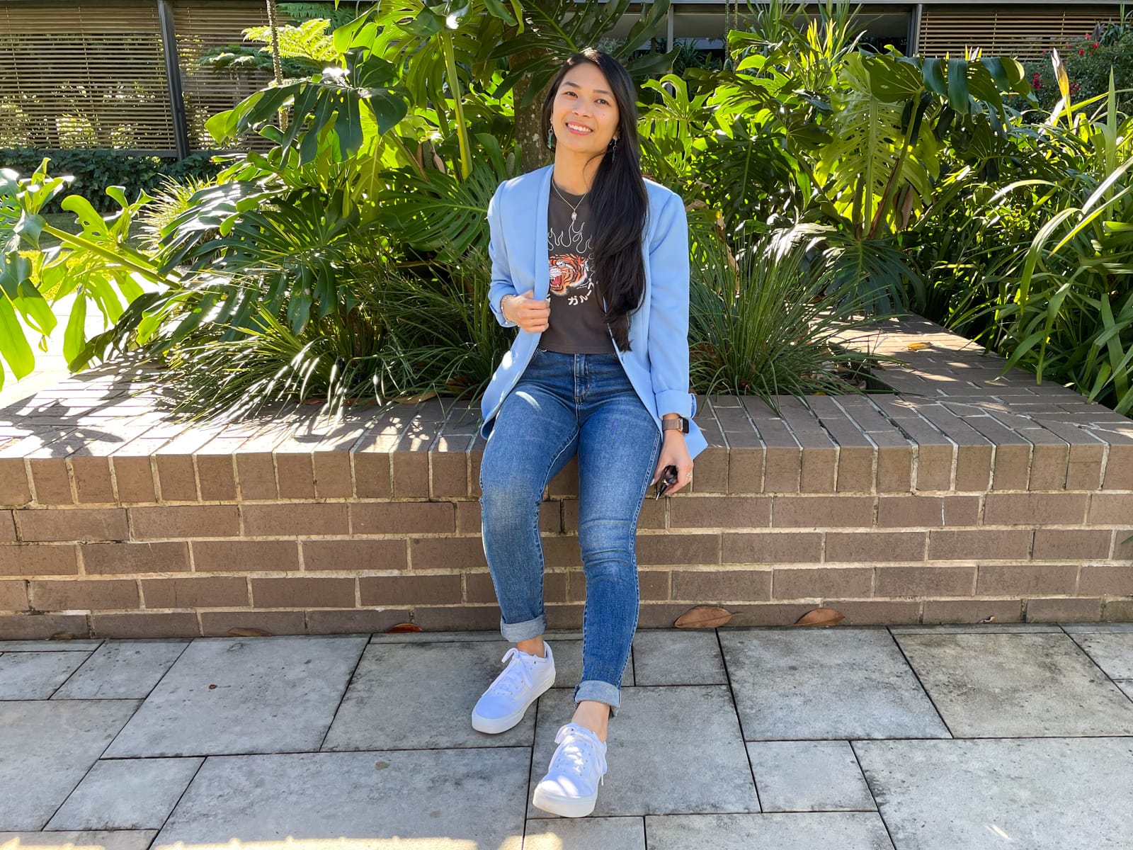 An Asian woman with dark hair, sitting on a brick wall. She is wearing a blue blazer over a dark grey graphic tee and blue jeans. She is wearing white sneakers.