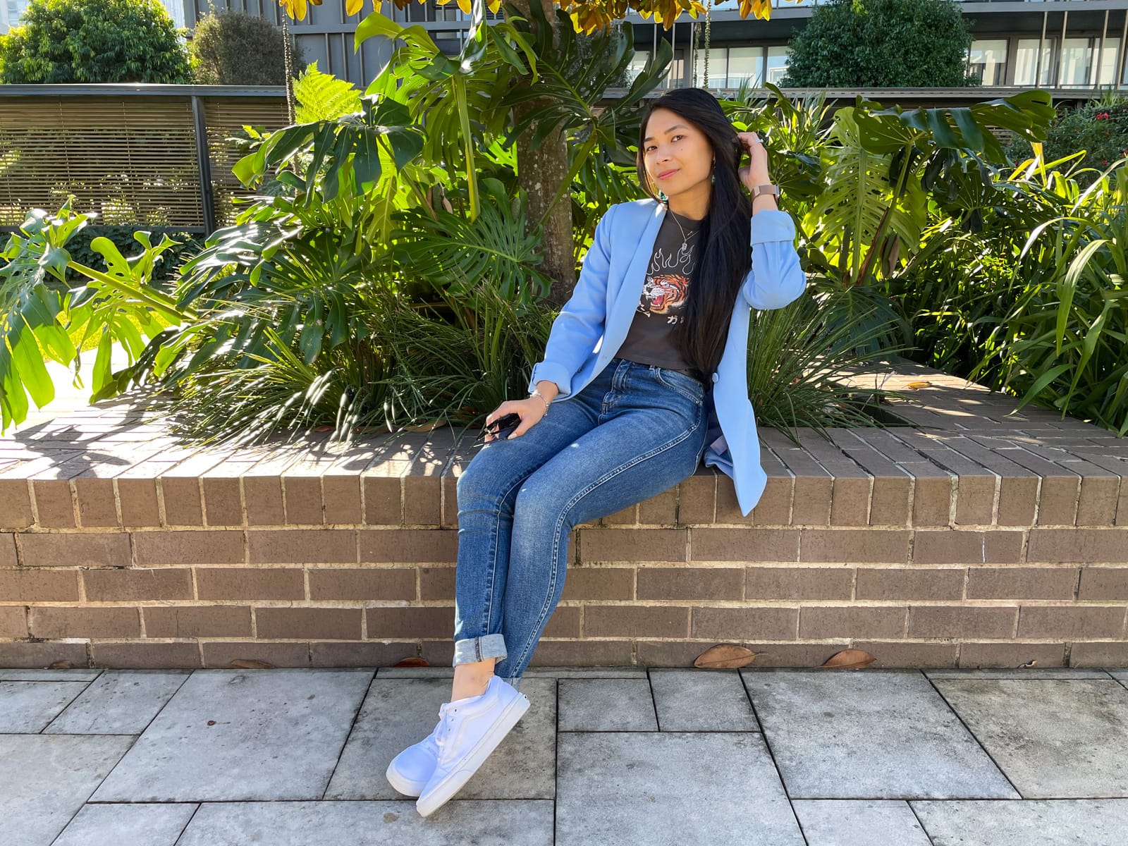 An Asian woman with dark hair, sitting on a brick wall with her knees bent and ankles crossed. She is wearing a blue blazer over a dark grey graphic tee and blue jeans. She is wearing white sneakers.