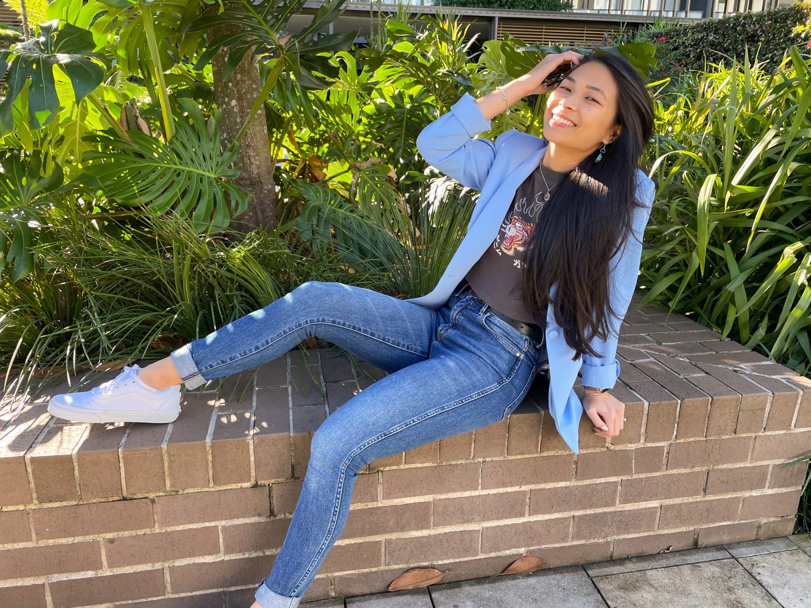 An Asian woman with dark hair, sitting on a brick wall with one leg bent up and a foot on the wall. She is wearing a blue blazer over a dark grey graphic tee and blue jeans. She is wearing white sneakers.