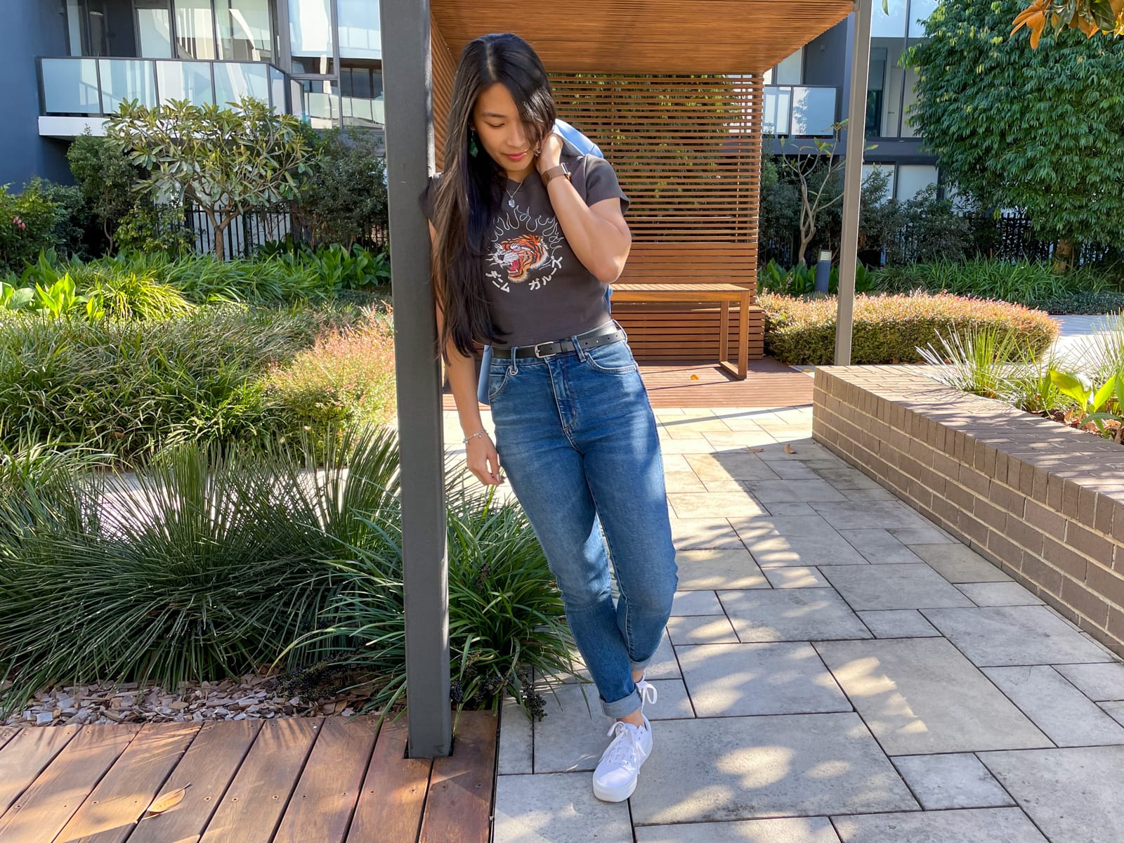 An Asian woman with dark hair, leaning against a square pole with her opposite hand over the back of her shoulder and carrying a blazer. She is wearing a dark grey graphic tee, blue jeans, and white sneakers.