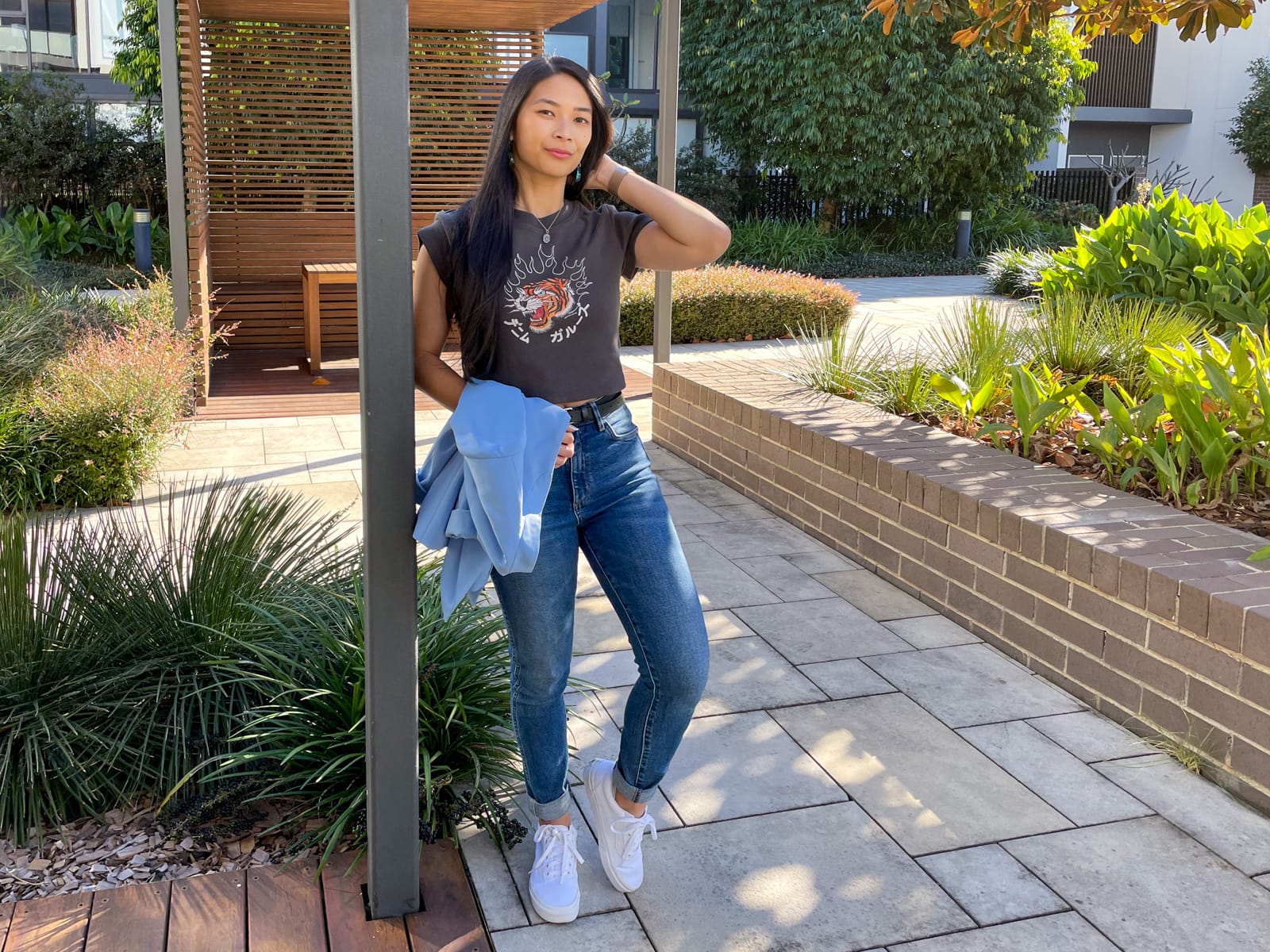 An Asian woman with dark hair, leaning against a square pole with one hand behind her head and the other holding a light blue blazer. She is wearing a dark grey graphic tee, blue jeans, and white sneakers.