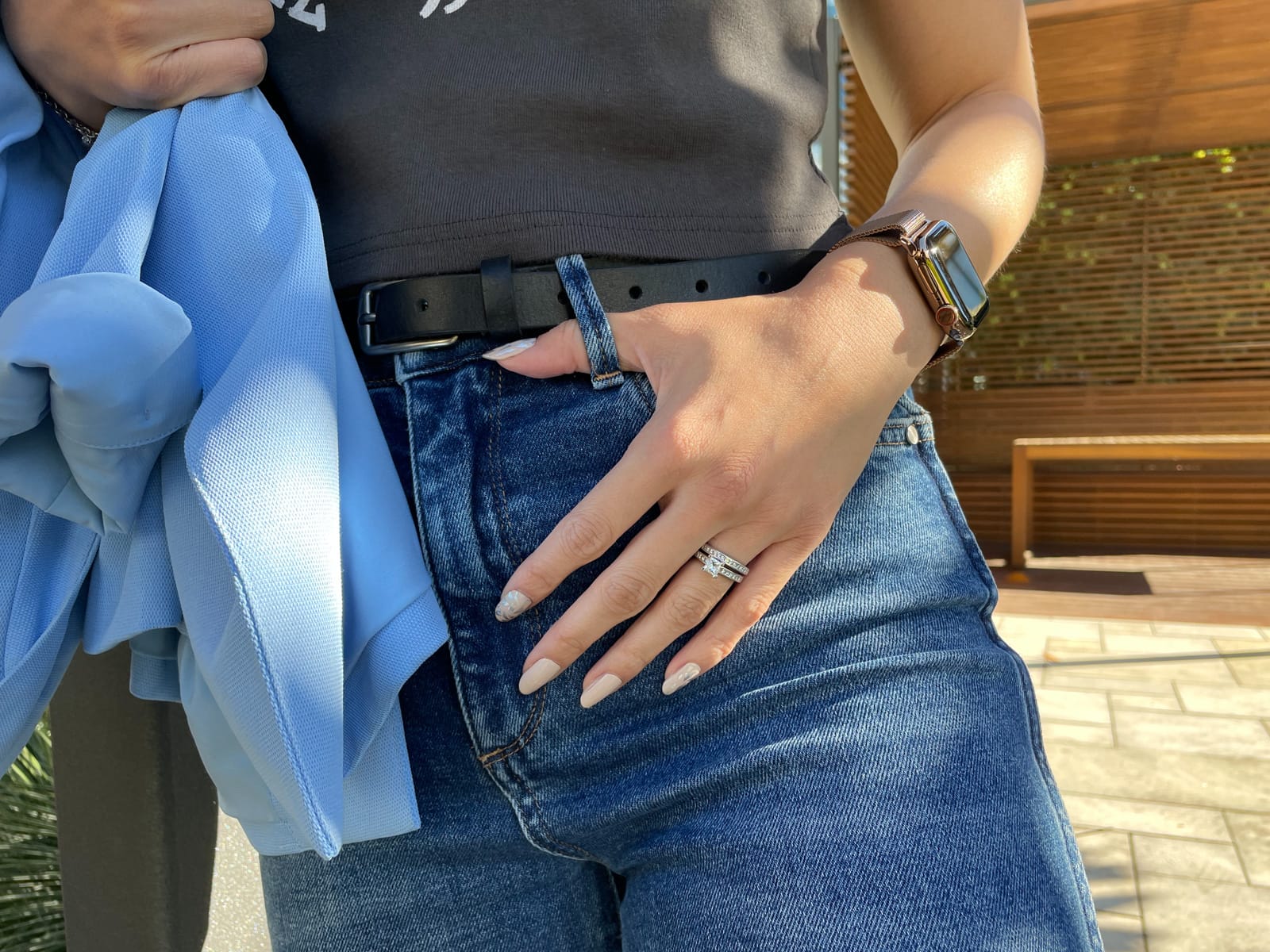 A close-up of a woman’s hand with her thumb through the belt loop on her blue jeans. She has a set of silver wedding rings on her fourth finger and her nails are light nude coloured with some silver accents. She is wearing a gold watch on her wrist.
