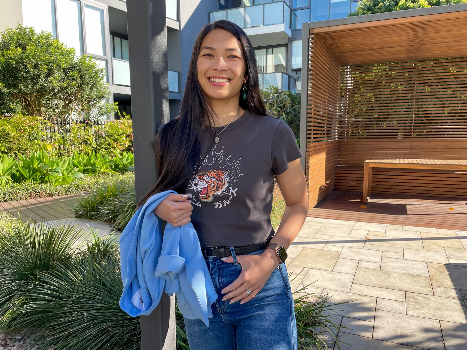 An Asian woman with dark hair, smiling. She is leaning against a square pole and has a thumb in one of the belt loops of her jeans. She is wearing a dark grey graphic tee and carrying a light blue blazer. In the background is a wooden cabana with a wooden table, some apartment buildings, and some planted trees.