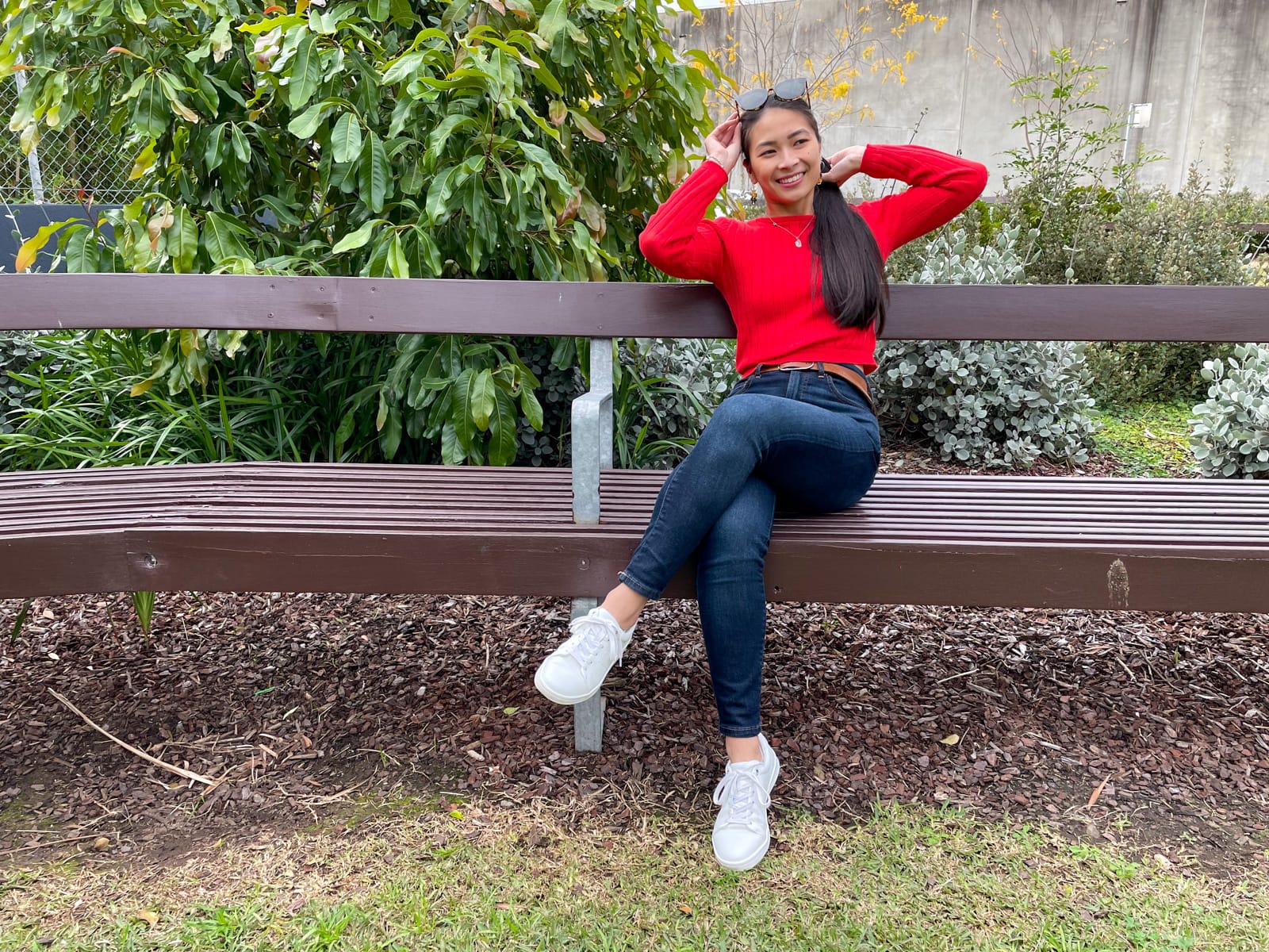 An Asian woman with long dark hair tied back in a low ponytail, sitting on a wooden park bench. She is wearing a red long-sleeved top and dark blue jeans, with white sneakers. She has her hands near her head and has tortoiseshell sunglasses on top of her head.