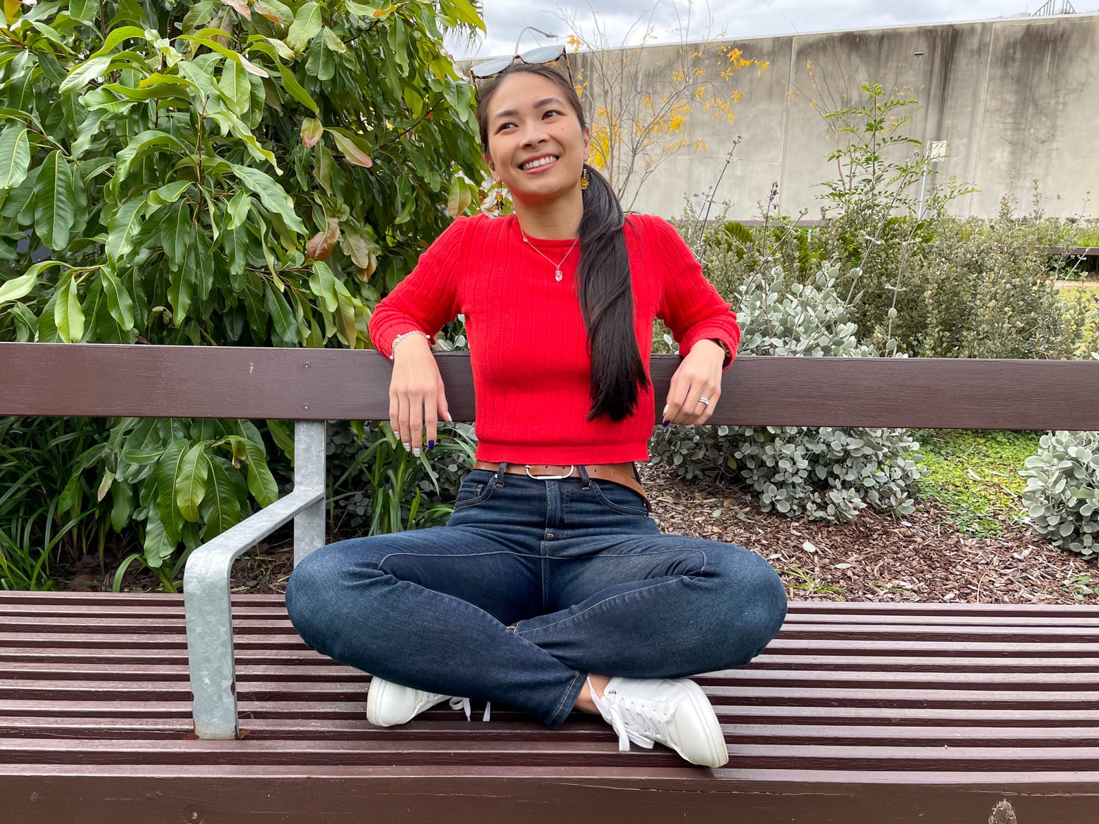 An Asian woman with long dark hair tied back in a low ponytail, sitting on a wooden park bench with her legs crossed. She is wearing a red long-sleeved top and dark blue jeans, with white sneakers. She has her elbows resting on the back of the bench.