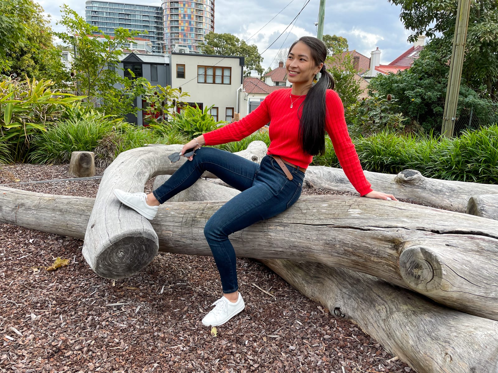 An Asian woman with long dark hair tied back in a low ponytail, sitting on a large tree log. She is wearing a red long-sleeved top and dark blue jeans, with white sneakers. She is leaning back slightly with one hand behind her.