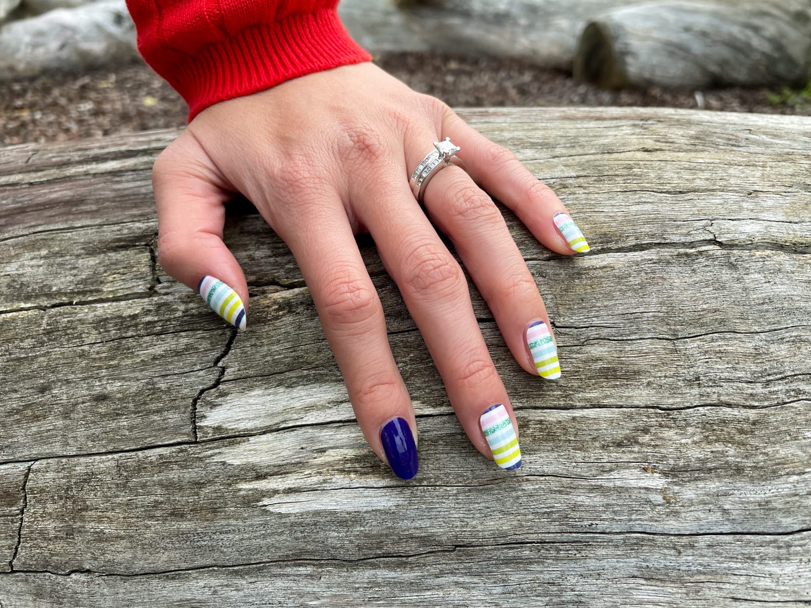 A close-up of a woman’s nail art which has colourful horizontal stripes and a blue accent nail on the index finder. There is a silver engagement ring and wedding ring on her ring finger. Her hand is resting on a giant log.