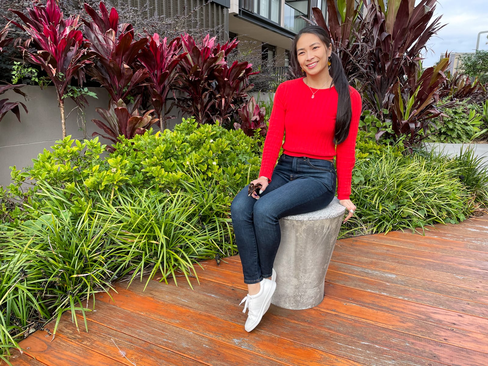 An Asian woman with long dark hair tied back in a low ponytail, sitting on a round concrete stool. She is wearing a red long-sleeved top and dark blue jeans, with white sneakers. Her ankles are crossed and she is holding a pair of sunglasses in her lap. There are bright green and red leafy plants in the background.