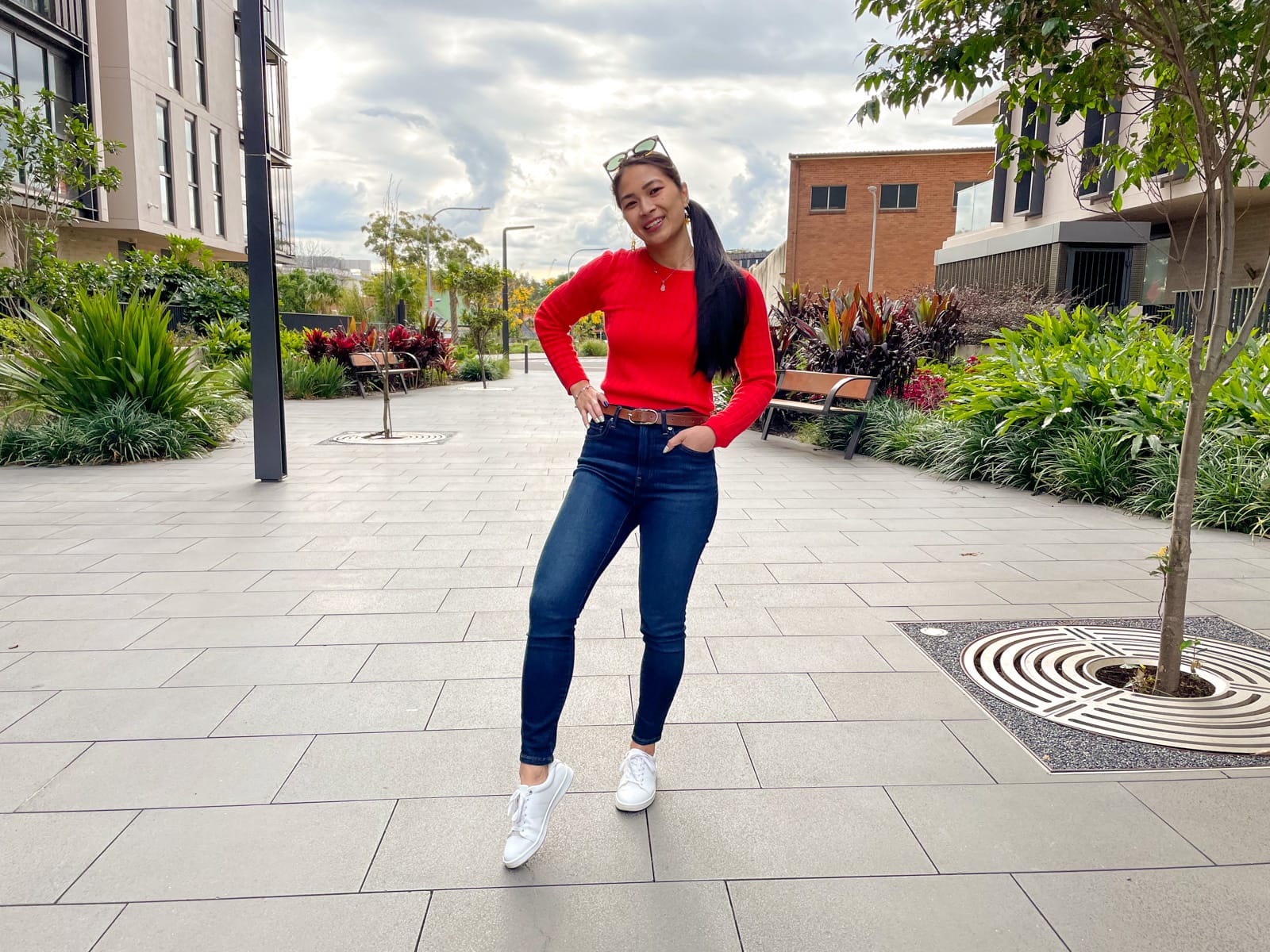 An Asian woman with long dark hair tied back in a low ponytail. She is wearing a red long-sleeved top and dark blue jeans, with white sneakers, and is standing on a concrete walkway with a hand in her pocket and another hand on her hip. She has sunglasses on top of her head.