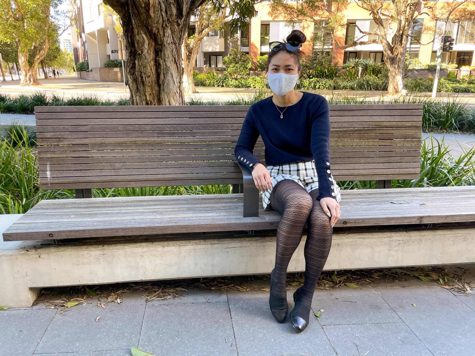 An Asian woman sitting on a wooden bench on a concrete path. She has dark hair tied back in a high bun. She is wearing a navy sweater with button details on the lower part of the sleeves, black and white checkered shorts, lace patterned tights and low profile black loafers. She is wearing a lace face covering over her nose and mouth, and has round sunglasses on top of her head. One of her elbows is resting on an armrest in the middle of the bench.