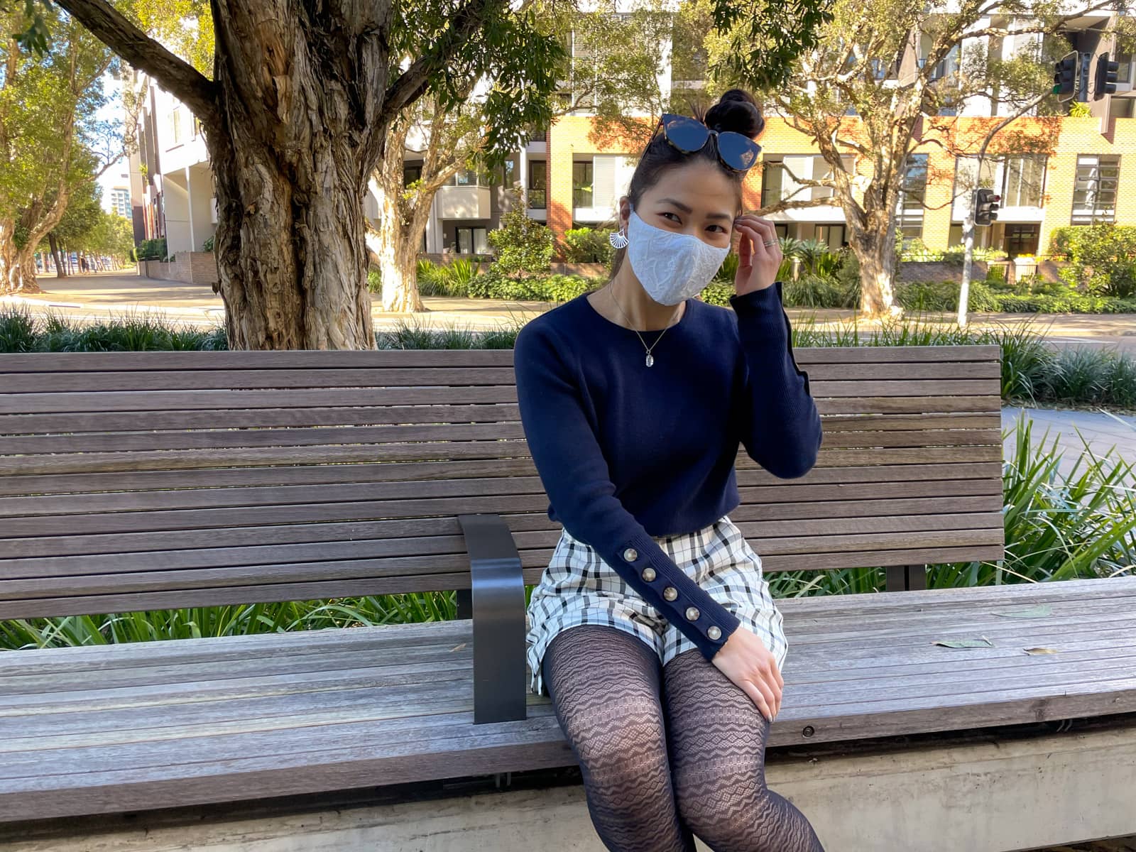 An Asian woman sitting on a wooden bench. She has dark hair tied back in a high bun. She is wearing a navy sweater with button details on the lower part of the sleeves, black and white checkered shorts, and lace patterned tights. She is wearing a lace face covering over her nose and mouth, and round sunglasses on top of her head. One of her arms is crossed over and resting on her opposite knee.