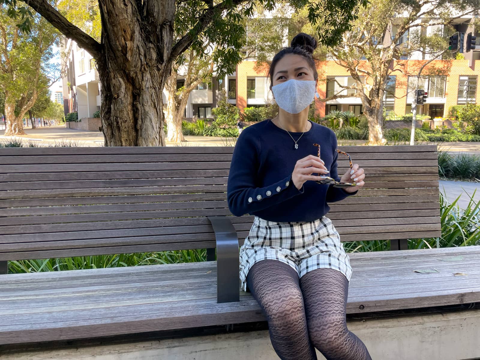 An Asian woman sitting on a wooden bench. She has dark hair tied back in a high bun. She is wearing a navy sweater with button details on the lower part of the sleeves, black and white checkered shorts, and lace patterned tights. She is wearing a lace face covering over her nose and mouth, and is holding a pair of round sunglasses in her hands.