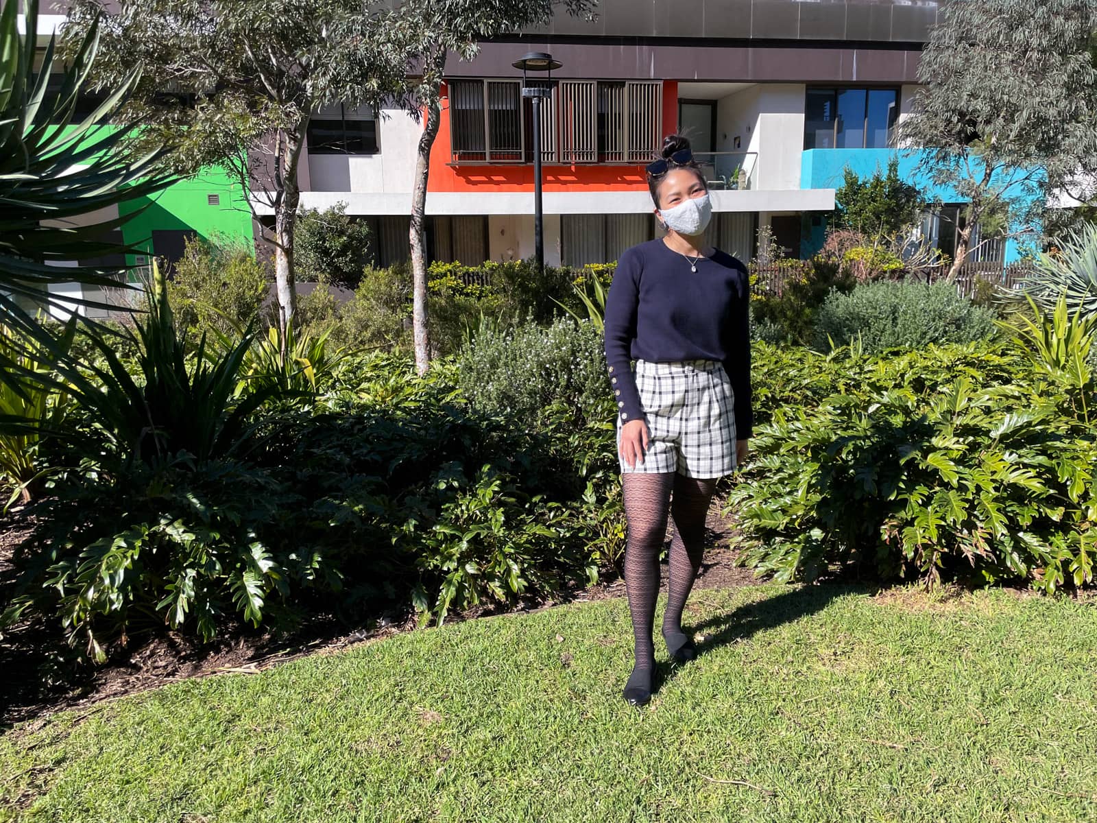 An Asian woman standing in a grassy park, with apartment buildings in the background. She has dark hair tied back in a high bun. She is wearing a navy sweater with button details on the lower part of the sleeves, black and white checkered shorts, and lace patterned tights. She is wearing a lace face covering over her nose and mouth.