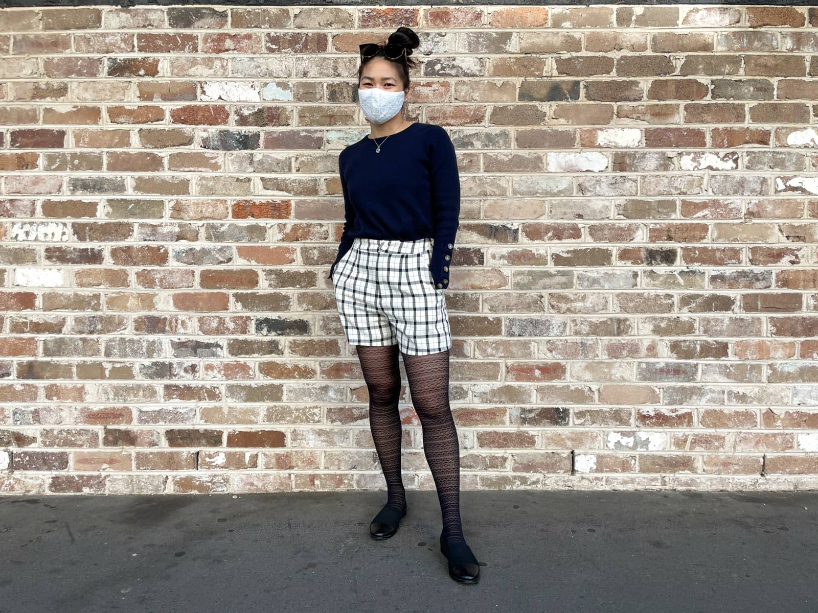 An Asian woman standing in front of a brown textured brick wall. She has dark hair tied back in a high bun. She is wearing a navy sweater with button details on the lower part of the sleeves, black and white checkered shorts, and lace patterned tights. She is wearing a lace face covering over her nose and mouth. Her hands are in her pockets.