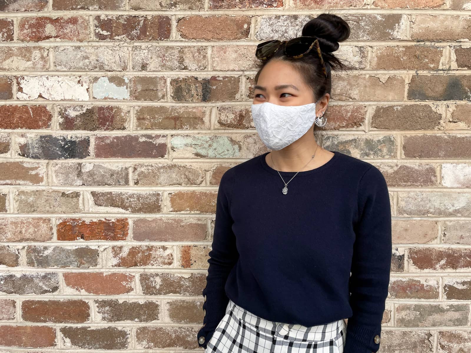 An Asian woman standing in front of a brown textured brick wall. She has dark hair tied back in a high bun. She is wearing a navy sweater and has her hands in her pockets as she looks away from the camera. She is wearing a white lace face covering over her nose and mouth. Her earrings are silver with the earrings in her bottom lobes being silver and fan-shaped.