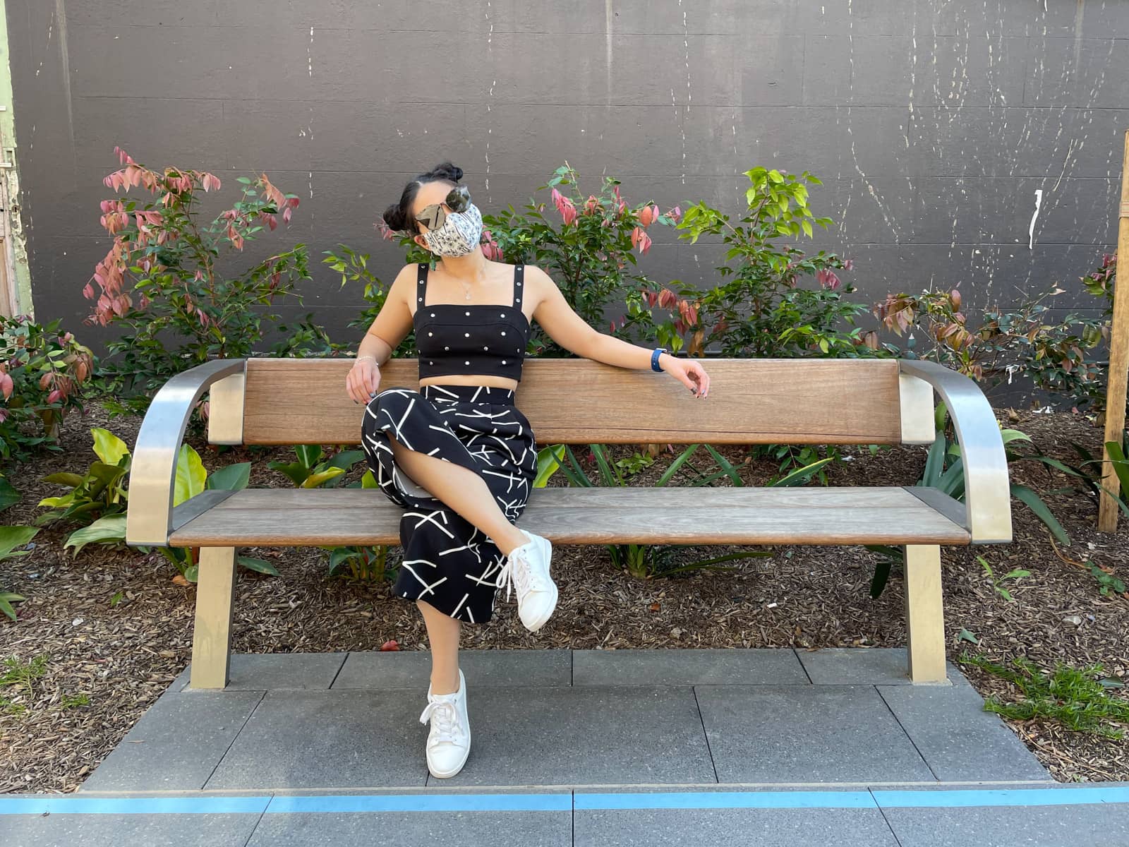 image 7: Same woman and outfit as image 1, same setting as image 1. She is sitting outside, on a wooden bench with metal armrests. She has round tortoiseshell sunglasses covering her eyes. One leg is loosely crossed over the other and her elbows are resting on the back of the bench.