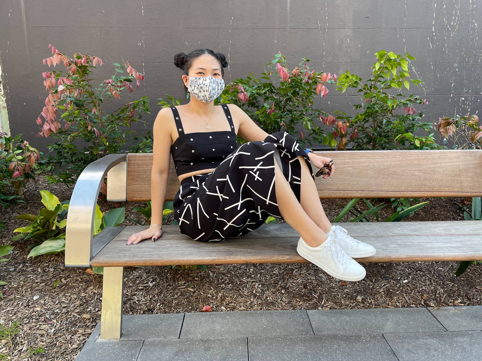 image 1: An Asian woman with dark hair, wearing a sleeveless black crop top. with round metal studs, black wide-leg cropped pants with a geometric line print, and white sneakers. Her hair is in two buns on either side of her head. She is wearing a light face mask with Japanese style navy floral print. The woman is holding her sunglasses in her hand and her other hand is on a wooden bench. Her knees are bent and her feet are on the bench and to her side.