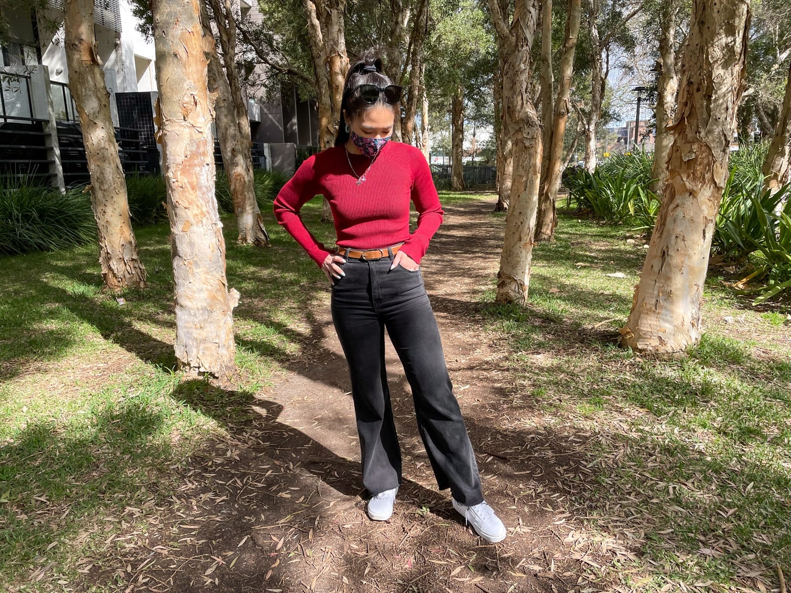 image 2: Same woman in the same outfit as image 1, standing on a dirt path between some spaced apart trees. She has white sneakers on. One of her hands is in her pocket and she is looking down, and has one foot slightly off the ground.