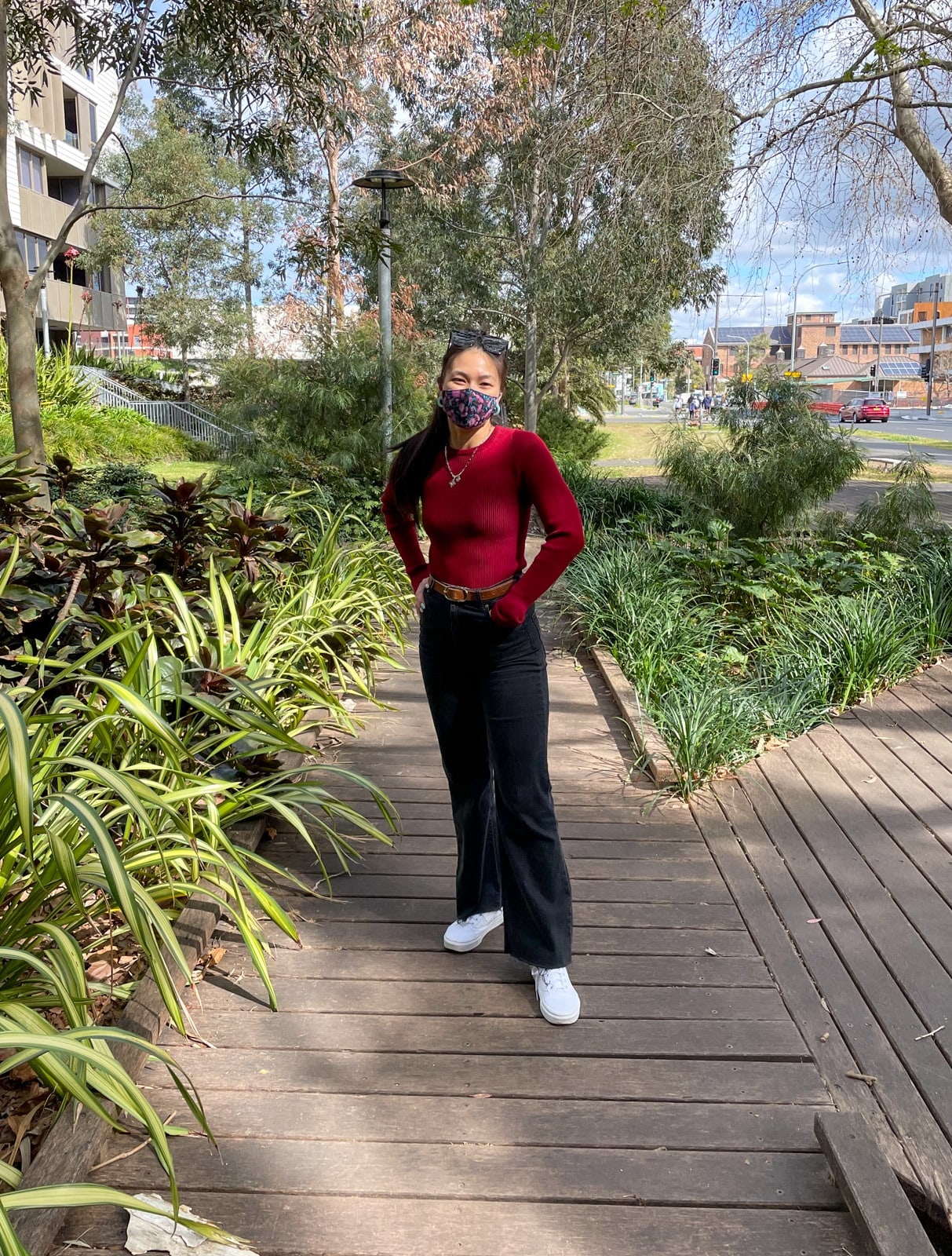 image 6: Same woman in the same outfit as image 1, and same setting. She has her hands in her jeans pockets and her elbows are slightly bent. This is a full body shot so you can see her white sneakers.