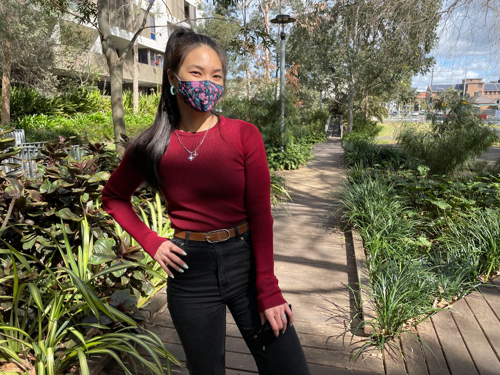 image 1: An Asian woman with dark hair in a ponytail. She is wearing a long sleeved red ribbed top, and black jeans with a tan belt. She has a deep and brightly patterned floral mask on. She has a hand on her hip and the other is holding a pair of sunglasses. She is standing on a wooden boardwalk with lots of greenery surrounding it.