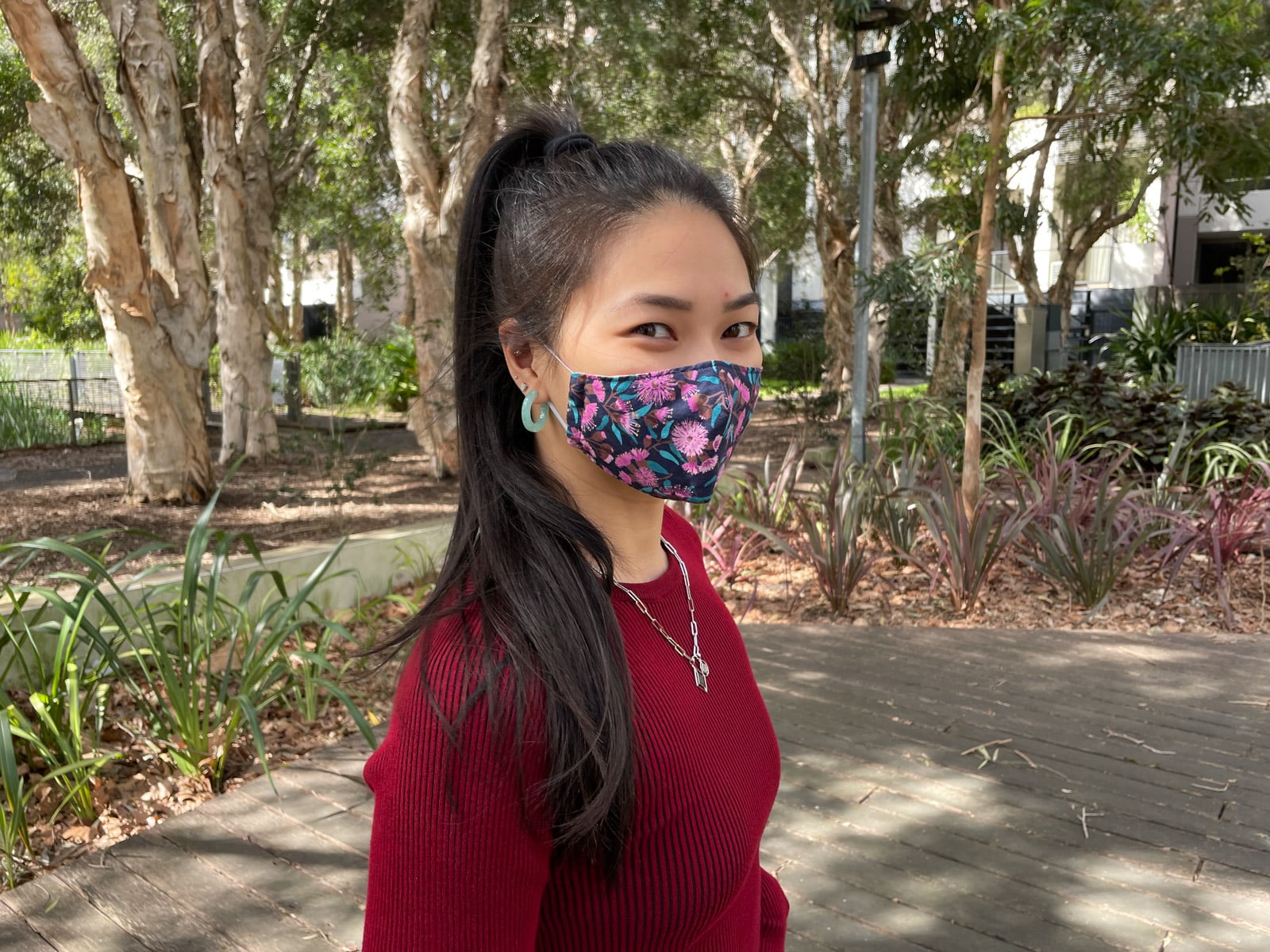 image 4: Same woman in the same outfit as image 1, but a close-up of her face, with her face angled slightly away from the camera. Her face mask is deep blue with pink and purple brightly coloured flowers on it. Her earrings are bright mint-coloured hoop earrings. The woman’s eyes indicate that she is smiling.