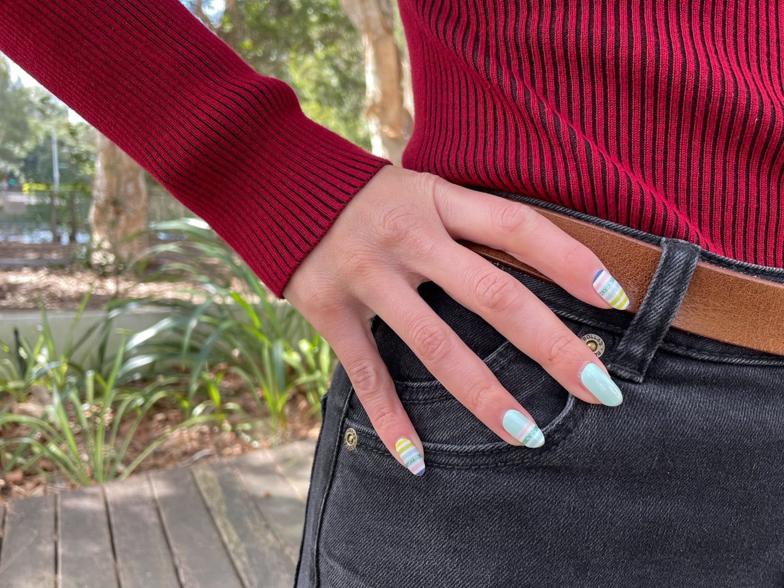 image 5: A close-up of the hand of the woman in image 1. Her hand is on her hip and her nails have multicoloured striped nail art, as well as some mint-coloured nails.