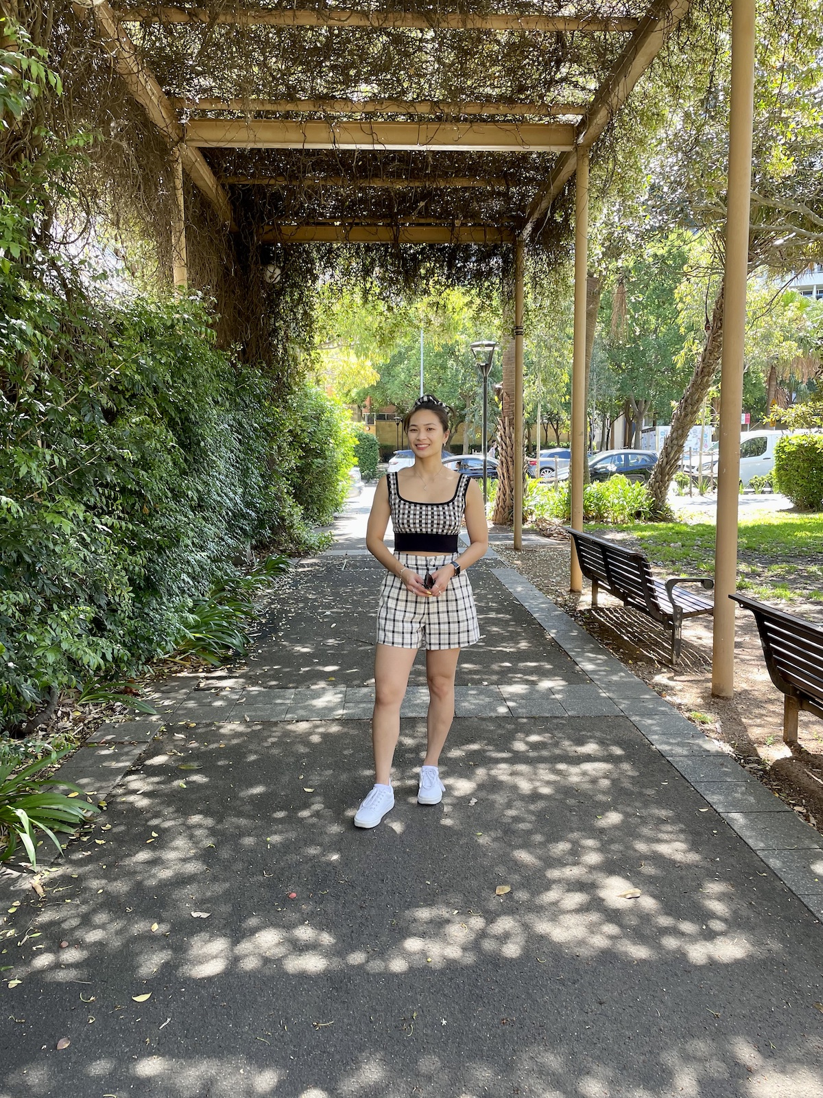 Image 1: An Asian woman with dark hair in a bun. She is standing under a sheltered path with vines and leaves growing over the wooden shelter. She is wearing a checkered top with black piping detail, white shorts with a black checkered pattern, and white sneakers. On the right are wooden park benches and on the left is green bush.