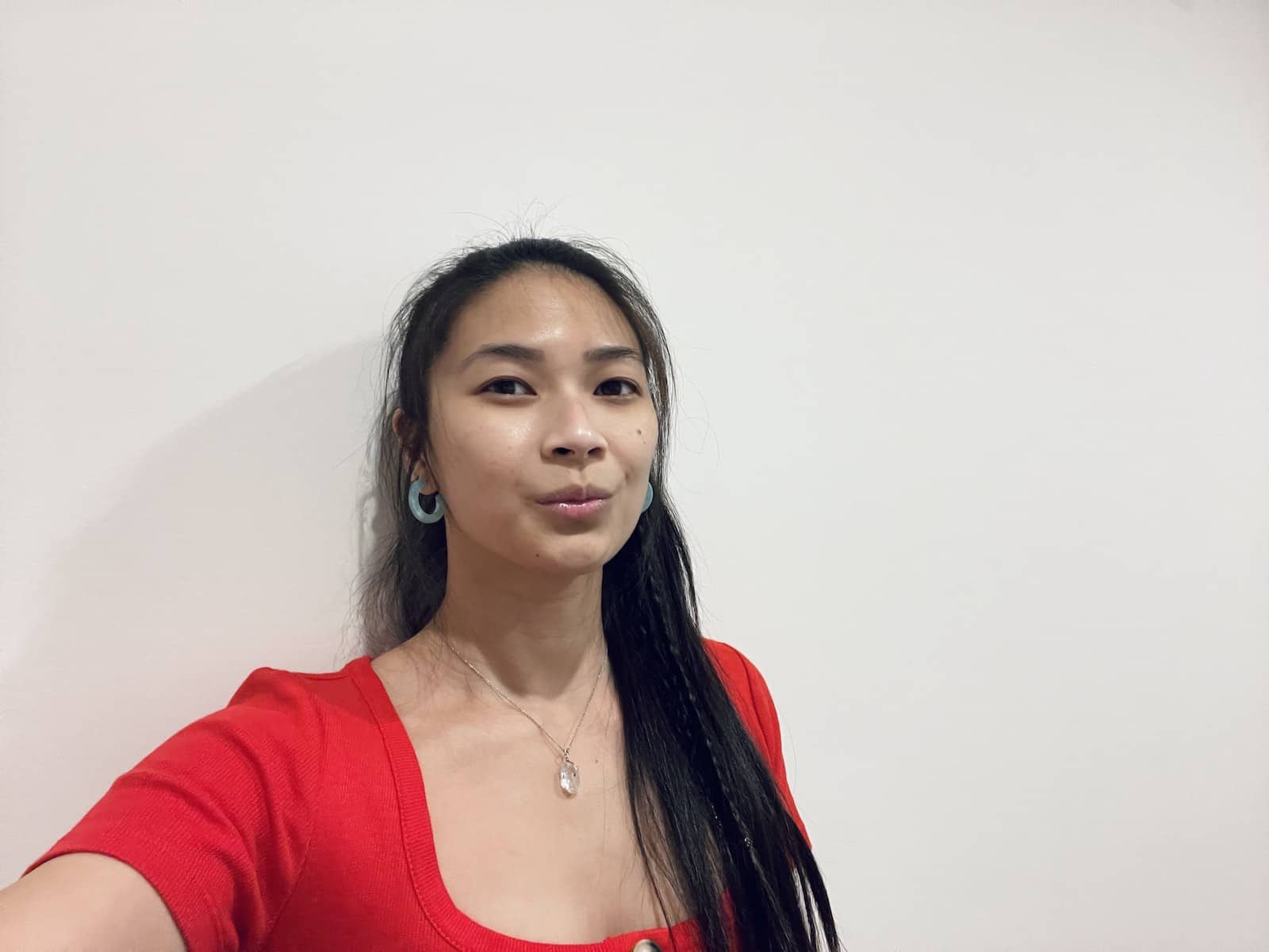 A selfie of an Asian woman with dark hair in a ponytail. She is wearing a red top with a button front. She has light blue resin hoop earrings and is also wearing a necklace with a herkimer diamond on the chain.
