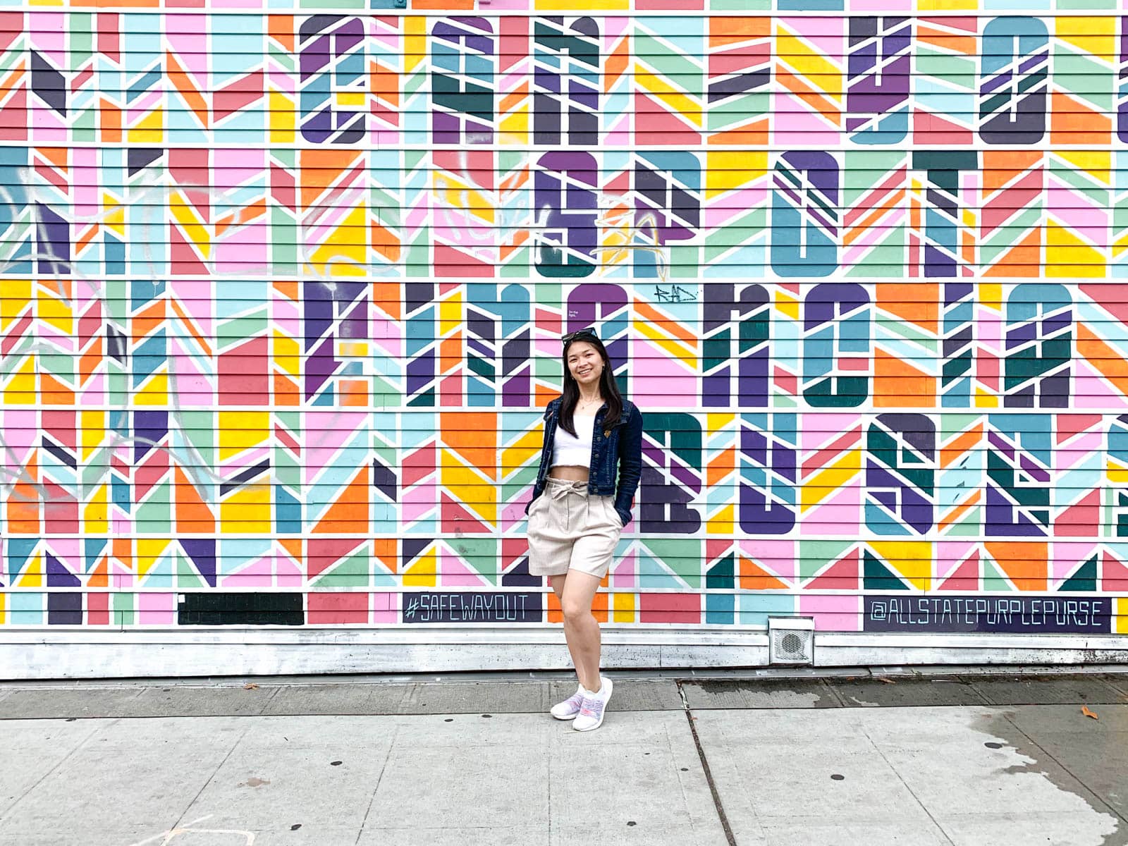 A woman with short dark hair, wearing a light coloured top and shorts, white shoes, and a denim jacket, standing in front of a colourful mural with her hands in her pockets.