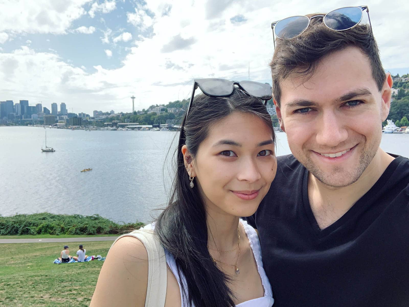 A selfie of a man and woman, with a large lake and a city skyline in the background. There is a couple sitting on the grass behind them. The man and woman have sunglasses on top of their heads and are smiling.