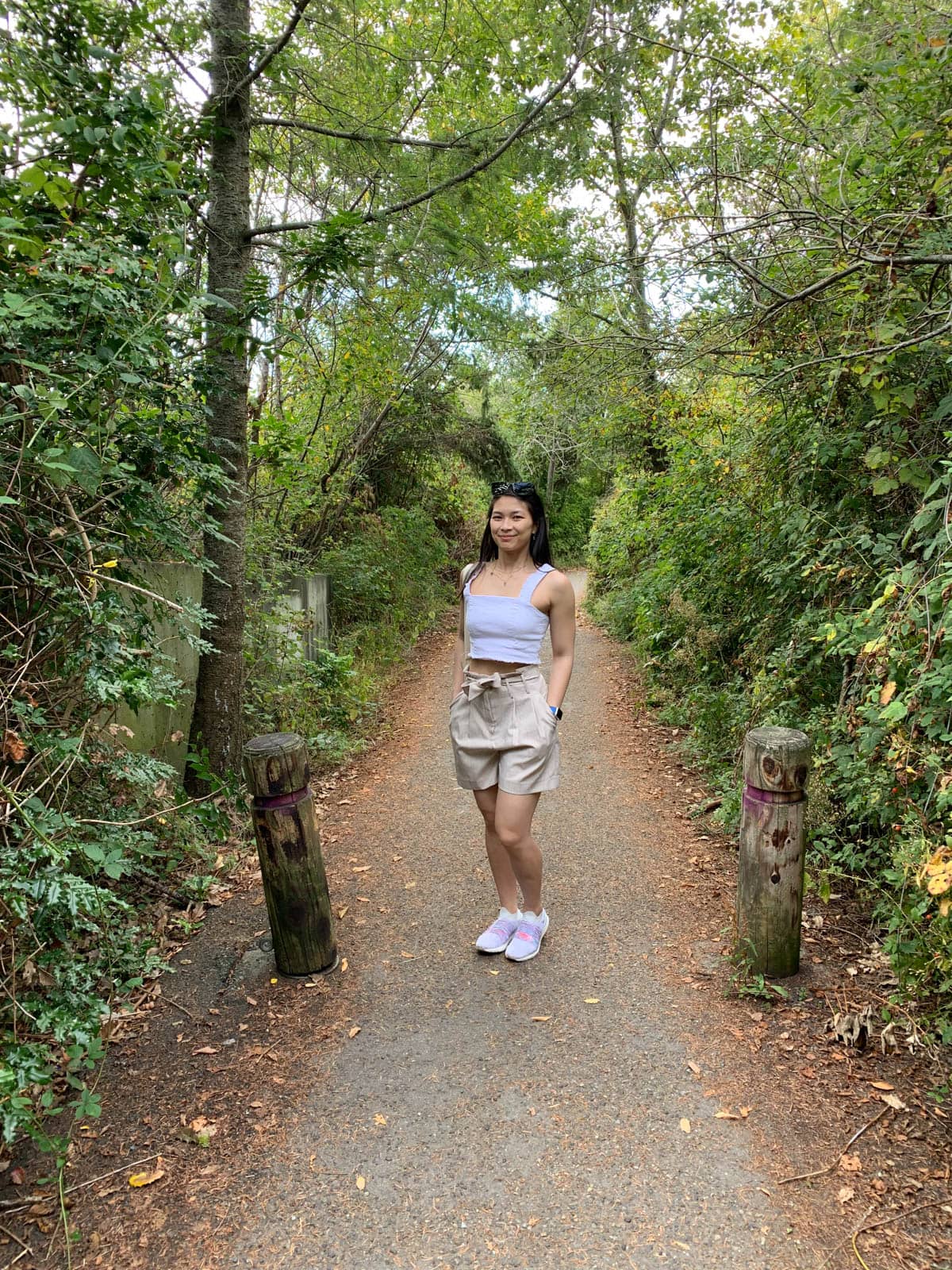 A woman with dark hair standing in the middle of a walking trail. Green shrubbery is visible on either side of her. She is wearing a white strapless top and natural coloured shorts, and has her hands in her pockets.