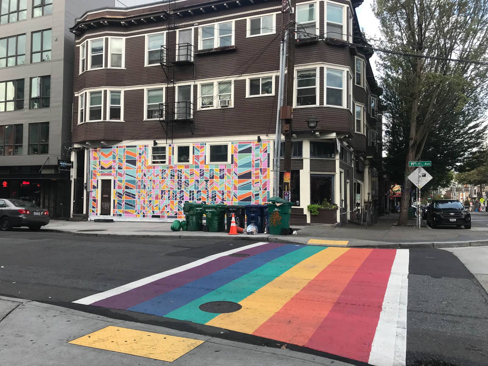 A rainbow-striped crosswalk painted on the road, with a colourful mural on the building on the opposite side of the road. The street is quiet.