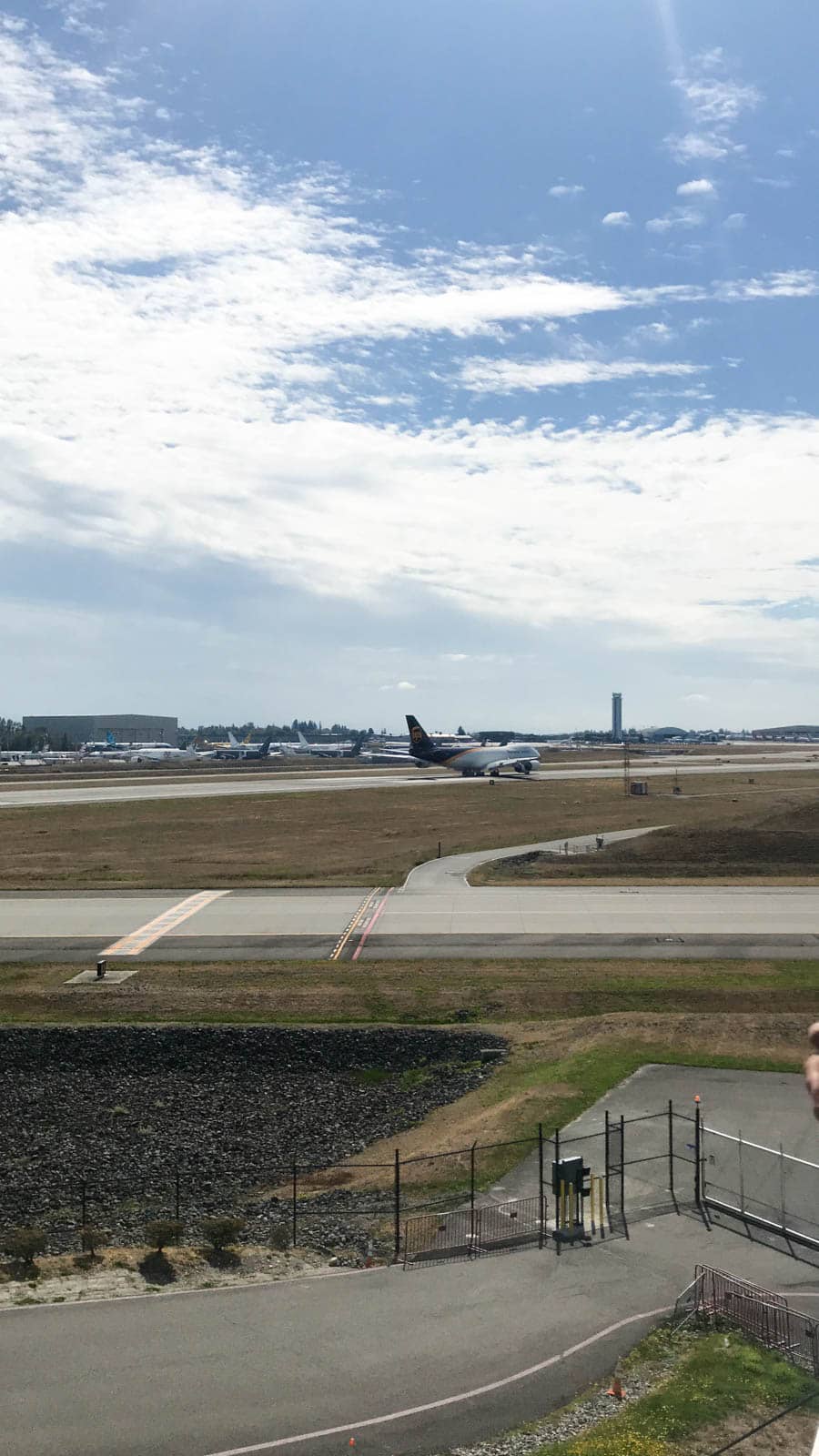 A plane on a runway in the distance. Further away, other planes can be seen. The skin is blue with some streaks of white clouds.