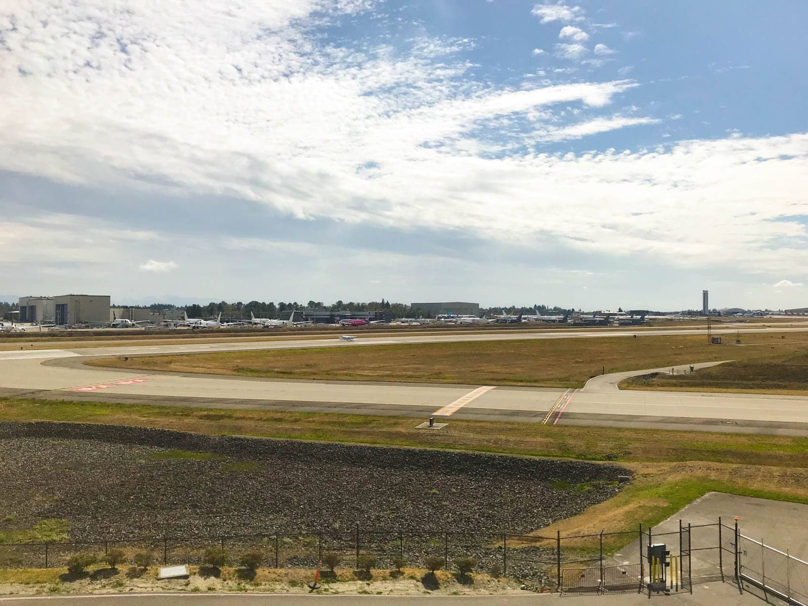 Empty runaways at the Boeing Factory in Seattle. The grass between the runways is a yellow-green colour. Some planes can be seen far off into the distance. The sky is blue with the clouds having created white streaks.