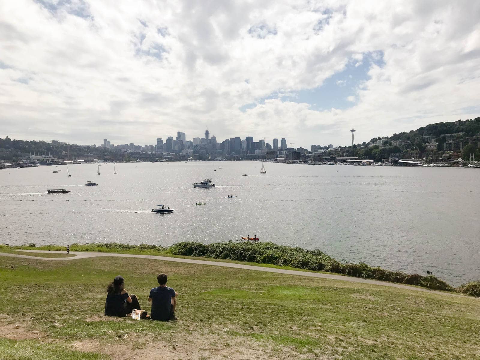 A view of Lake Union in Seattle, showing the Space Needle building prominent on the skyline. The sky is full of streaky clouds but it is a bright day. A couple sits in the foreground on the grass, looking out at the water.