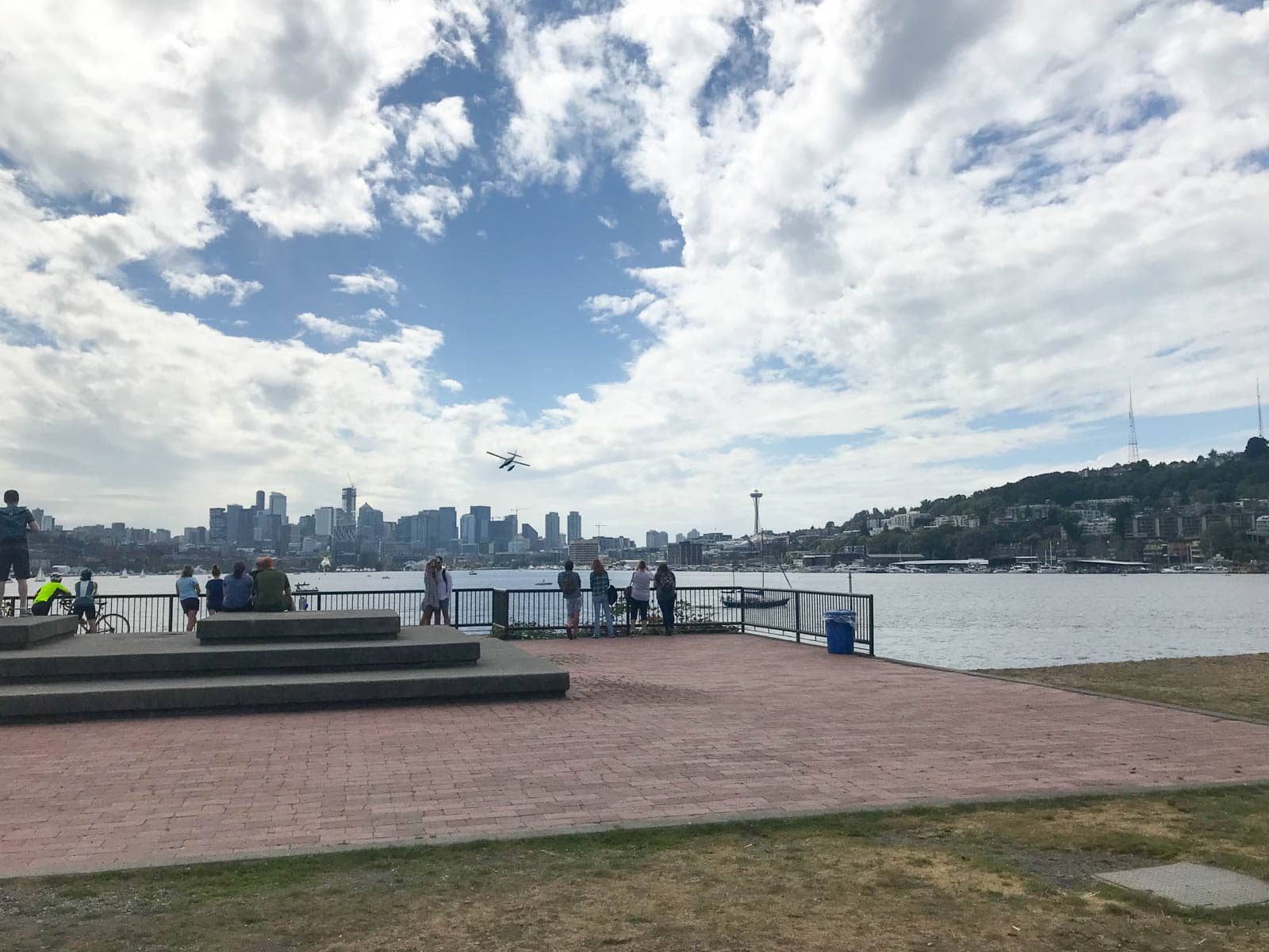 The city of Seattle seen from a viewing platform, with Lake Union in the foreground and the city skyline in the background. A seaplane overhead is heading into the distance. The sky is blue but streaked with clouds.