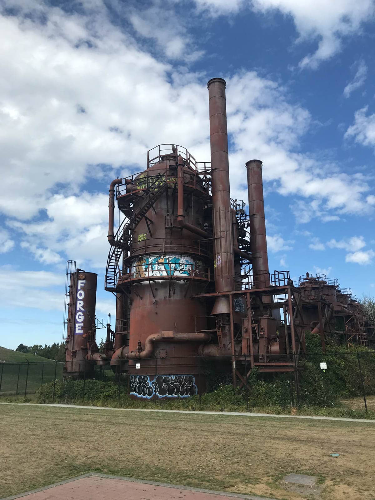 A large copper-brown-coloured structure, part of a coal plant with pipes and ladders. Some graffiti is on the structure. The sky is blue with some clouds.
